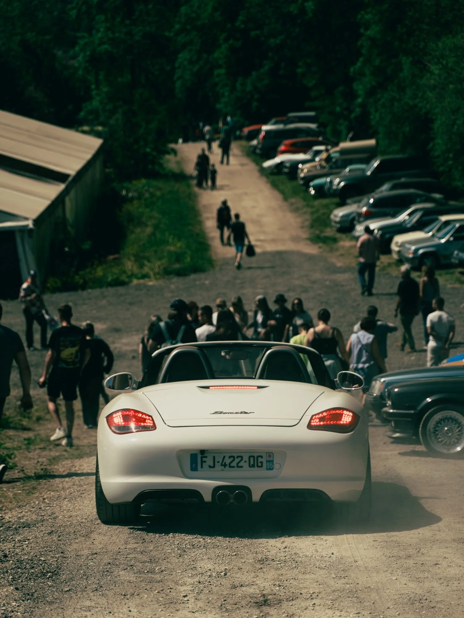 Une voiture blanche Porsche vue de l'arrière sur un chemin de terre, avec une foule de personnes et des voitures garées le long du côté droit, dans un environnement verdoyant.