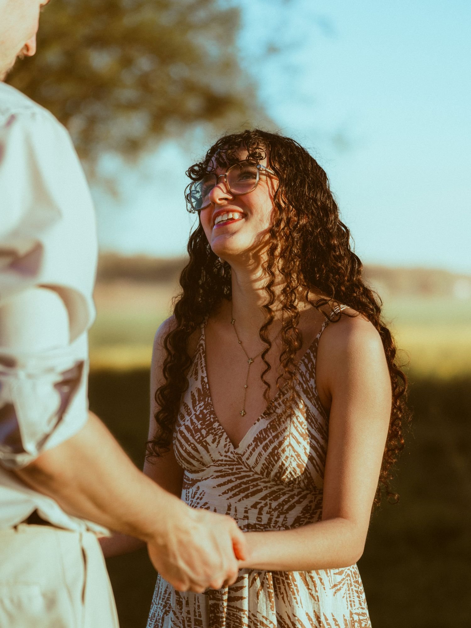 Une jeune femme souriante, portant une robe à motifs, serre la main d'une autre personne lors d'une rencontre en plein air sous un arbre.
