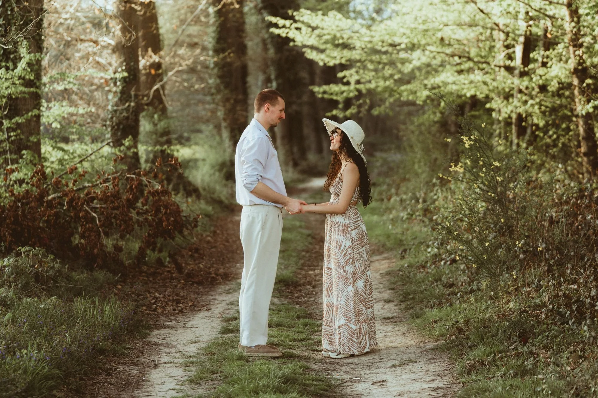 Un couple marié se tient la main dans un chemin forestier, le matin ou en fin d'après-midi, entouré d'arbres verts, dans une ambiance romantique et naturelle.