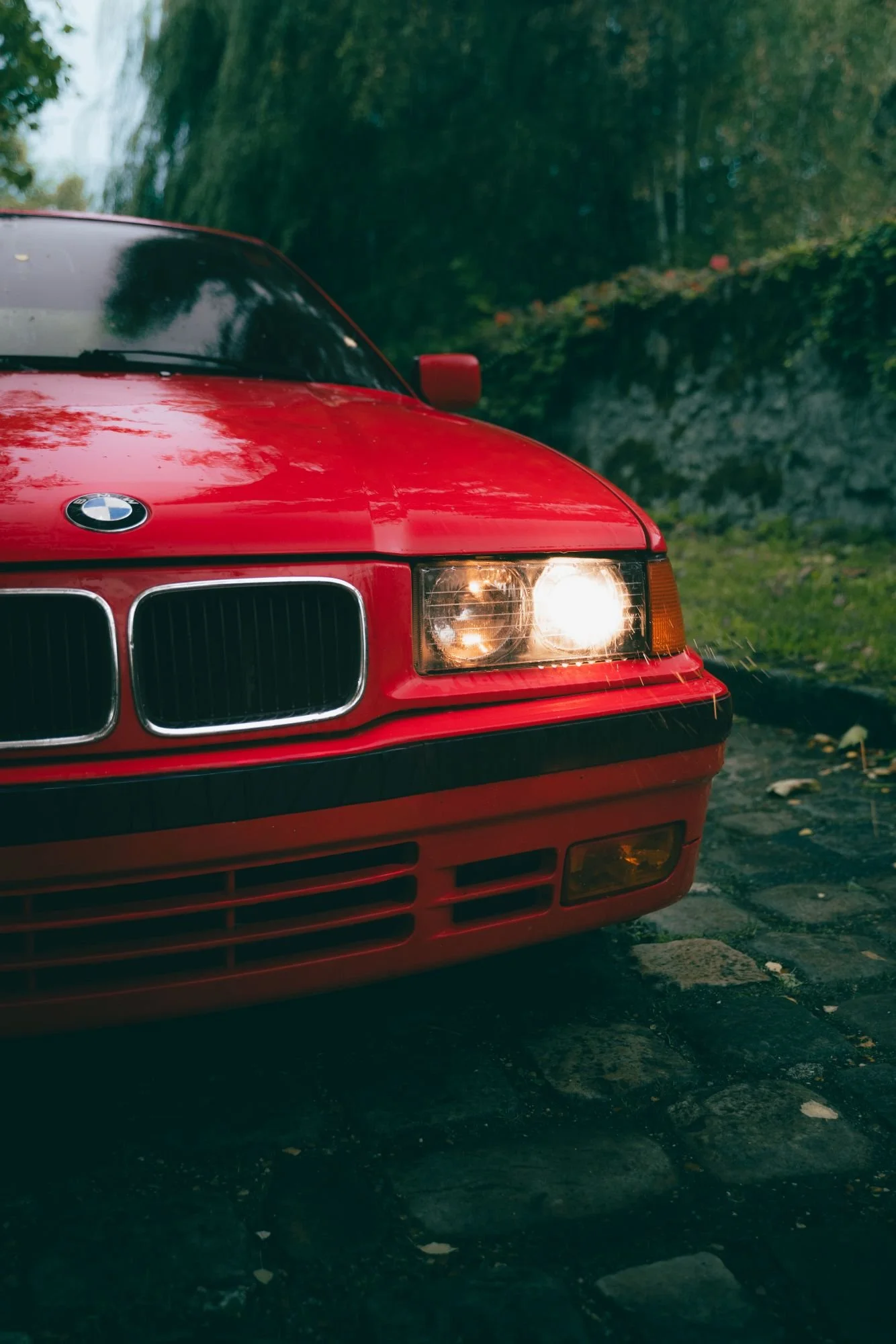 Une voiture rouge BMW stationnée sur une voie pavée, avec des arbres en arrière-plan, sous un ciel nuageux.