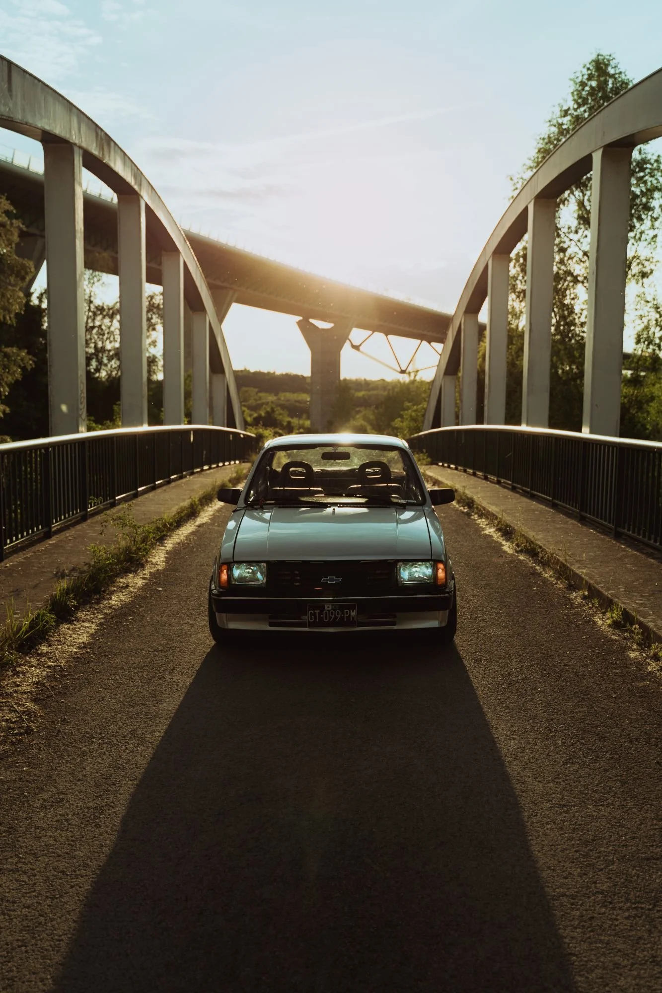 Une voiture noire garée sur un pont en fin d'après-midi, avec le soleil couchant derrière, entouré d'arbres et d'une structure de pont en arrière-plan.