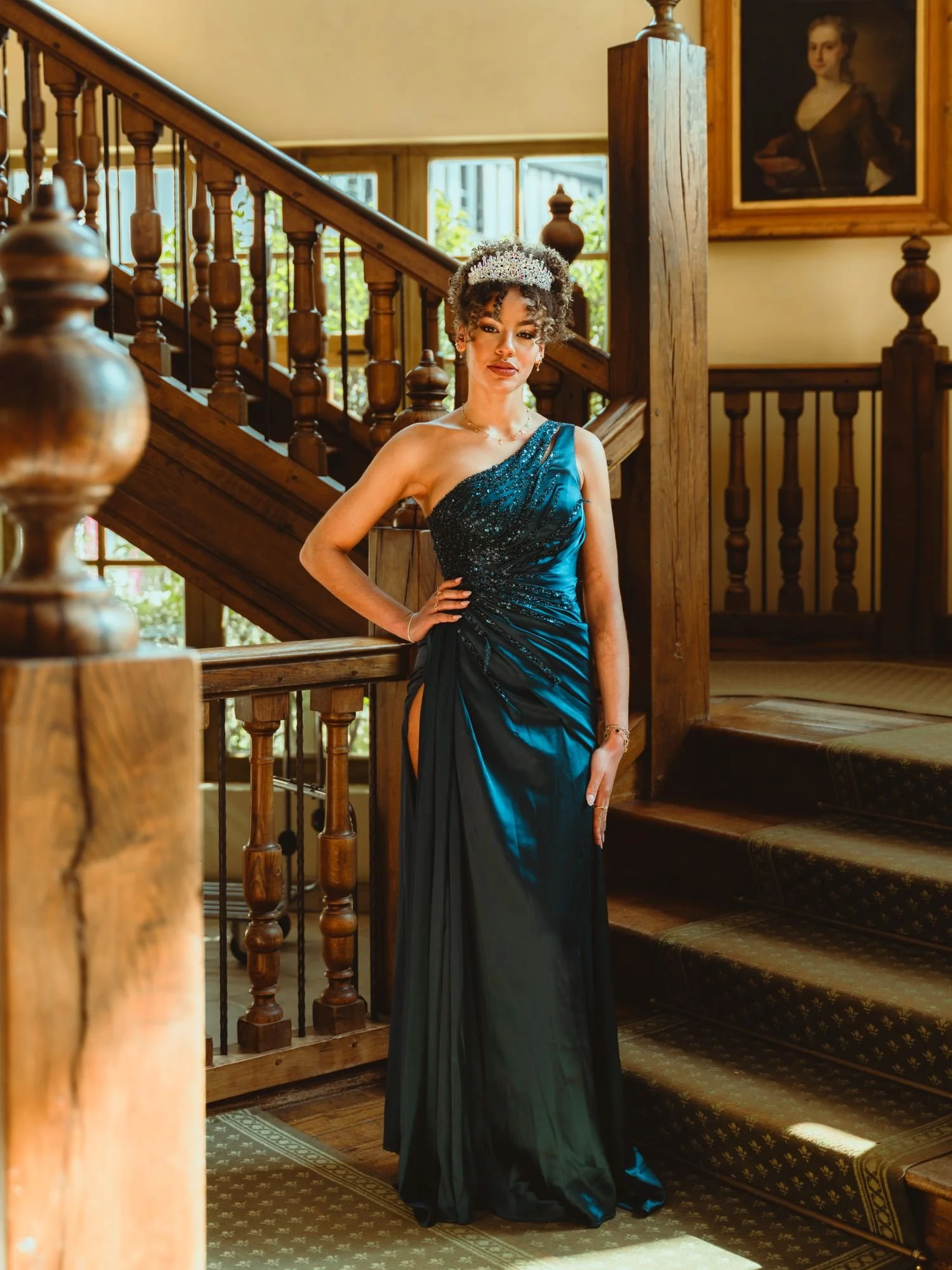 Jeune femme en robe de soirée bleue et noire, portant une couronne de fleurs, posant dans un intérieur en bois avec escalier en bois et tableau ancien au mur.