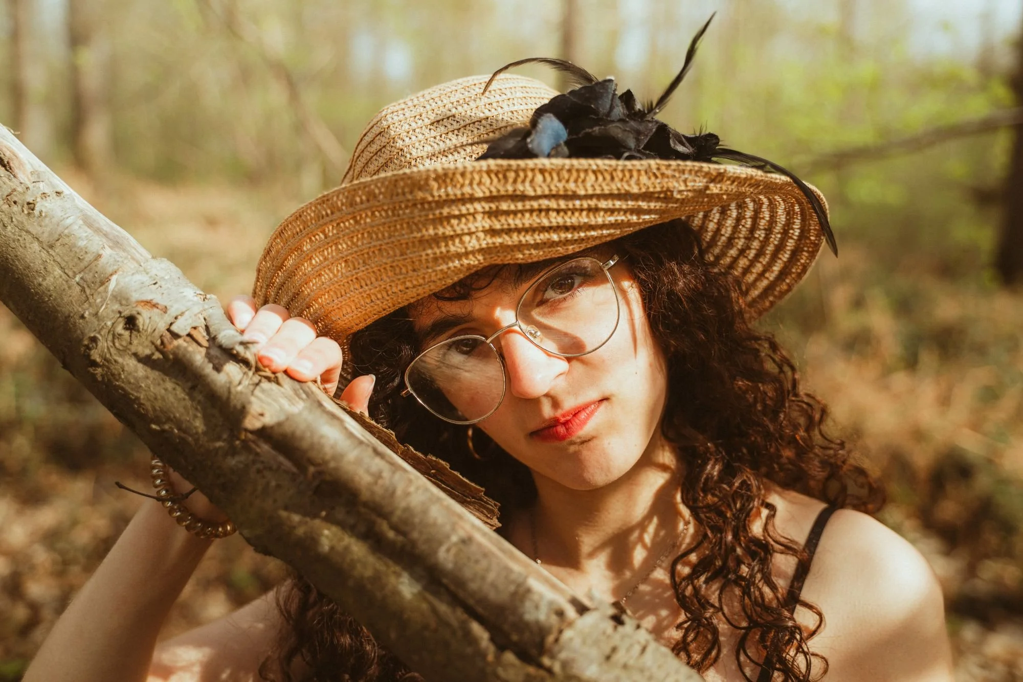 Une femme avec des cheveux bouclés, portant des lunettes et un chapeau en paille, regarde vers l'objectif tout en tenant un arbre dans une forêt ensoleillée.
