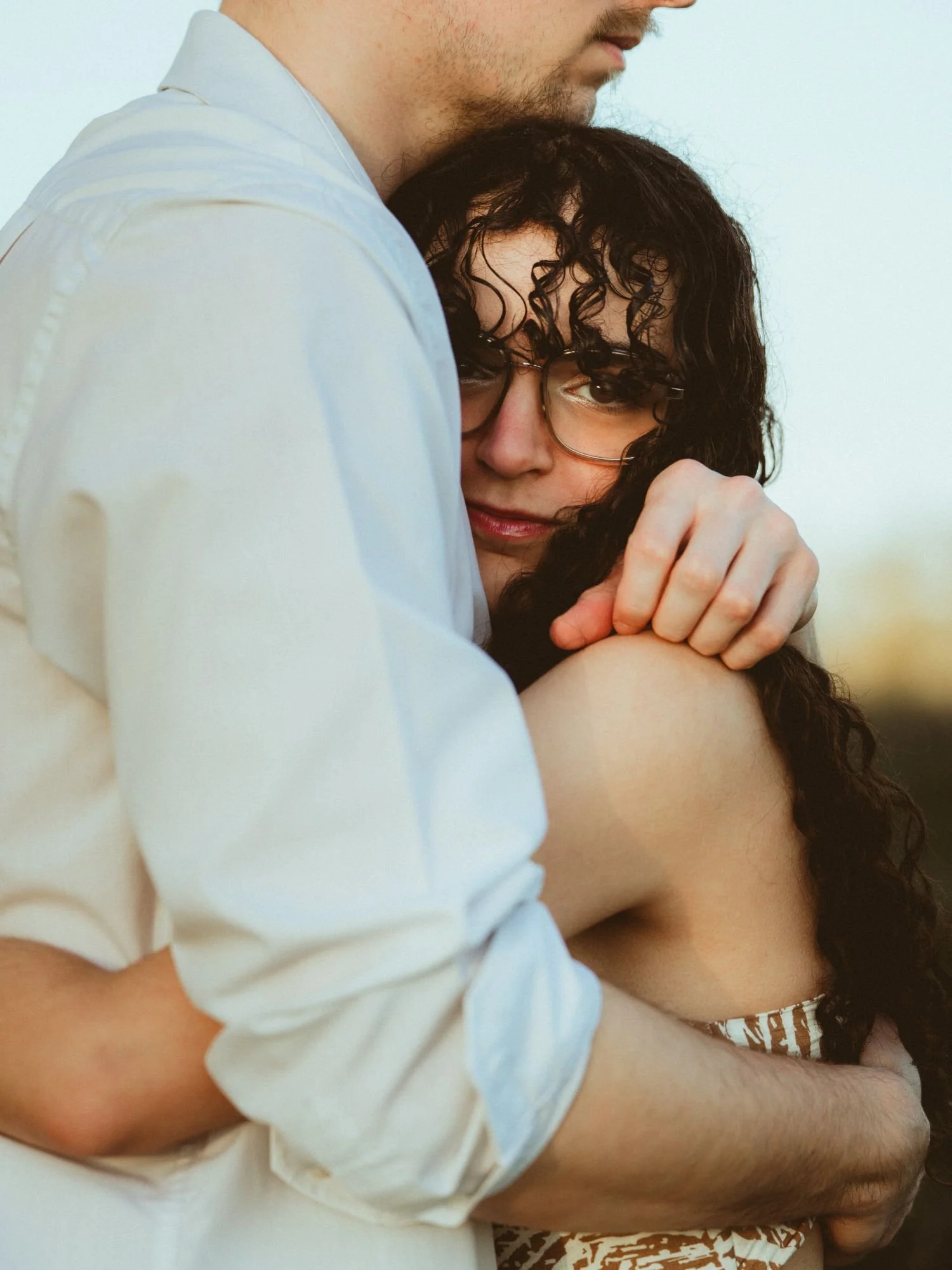 Un couple s'embrasse tendrement, l'homme porte une chemise blanche et la femme porte des lunettes, avec un coucher de soleil en fond.