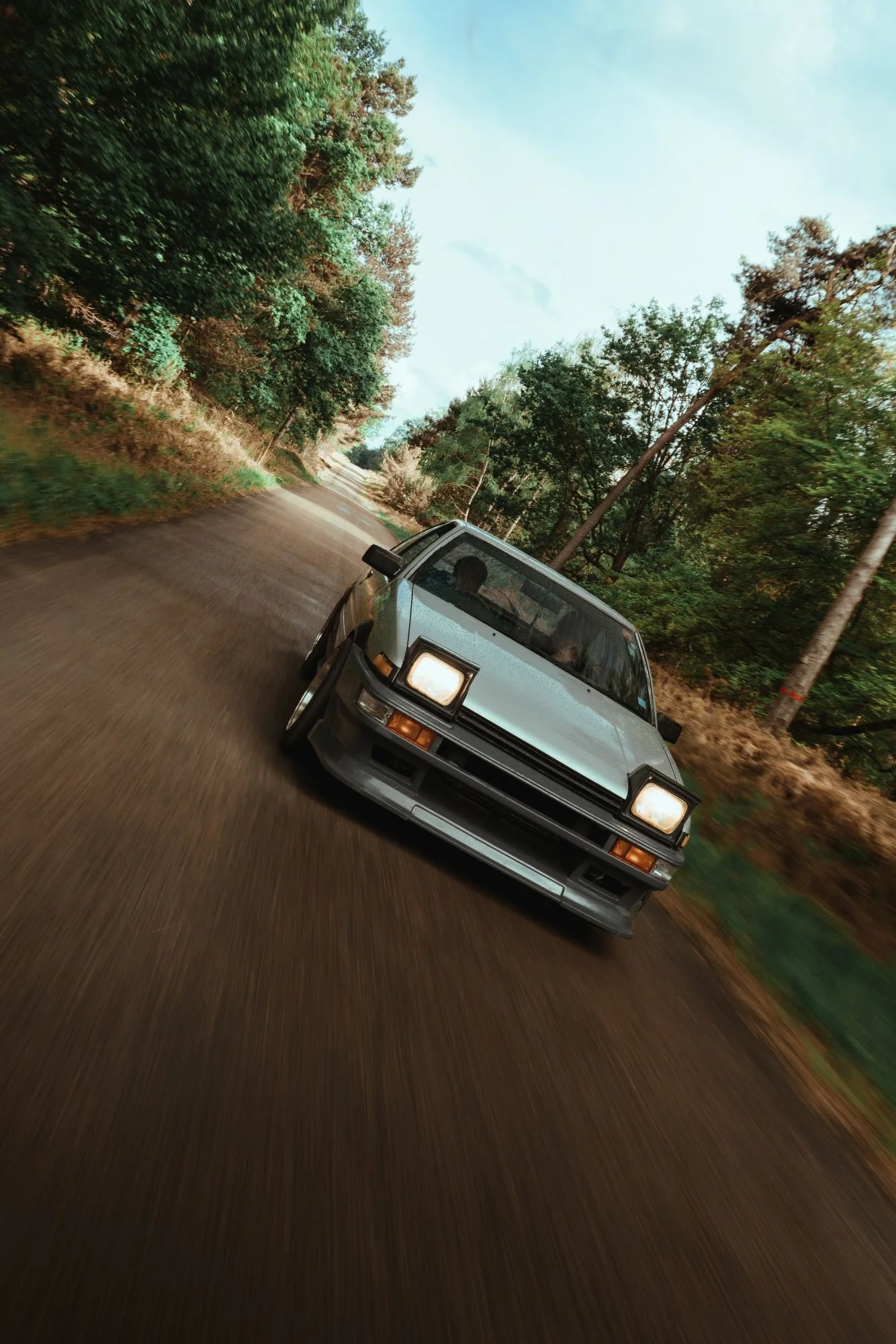 Voiture noire roulant sur une route forestière avec des arbres verts de chaque côté, ciel clair en arrière-plan.