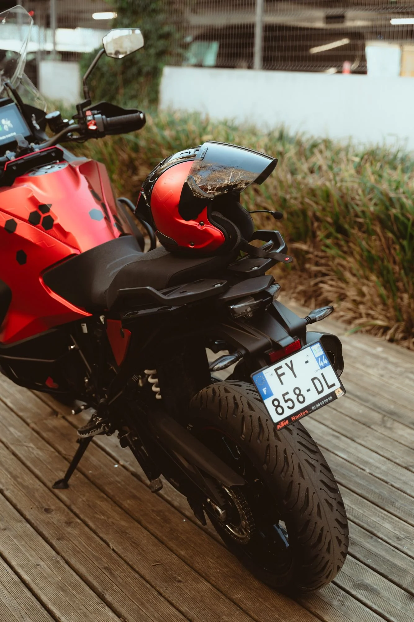 Motocyclette rouge avec casque rouge et noir posé sur la selle, stationnée sur une plateforme en bois, avec une casquette en visière réflective.