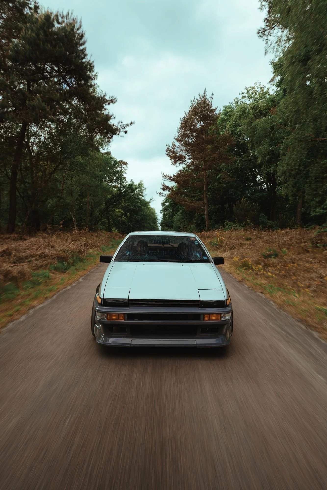 Une voiture blanche de style rétro se déplaçant sur une route bordée d'arbres, vue de face, en mouvement.