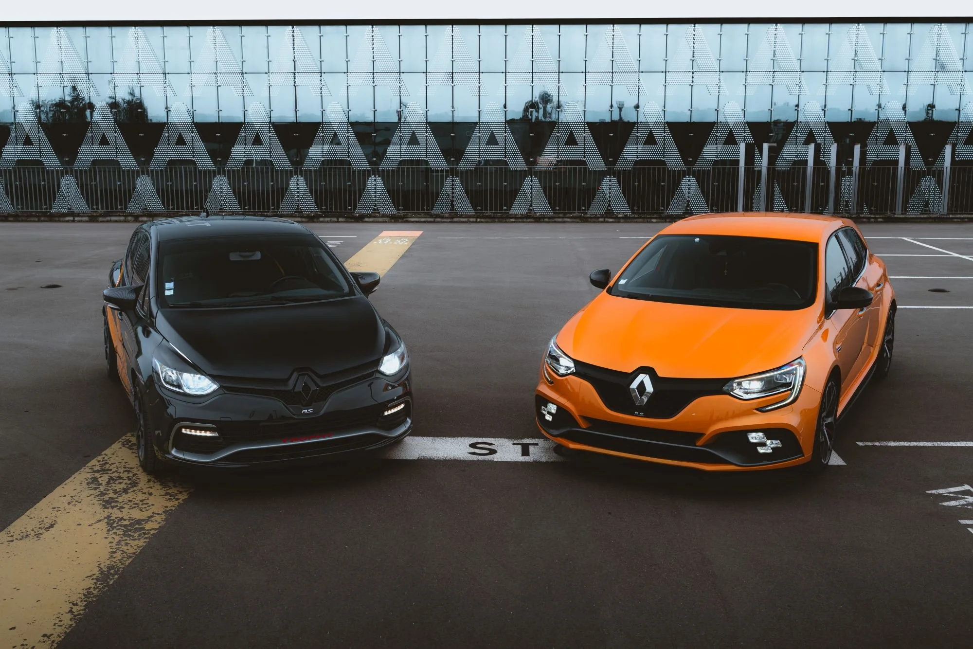 Deux voitures de marque Renault, une noire et une orange, stationnées côte à côte dans un parking en milieu urbain.