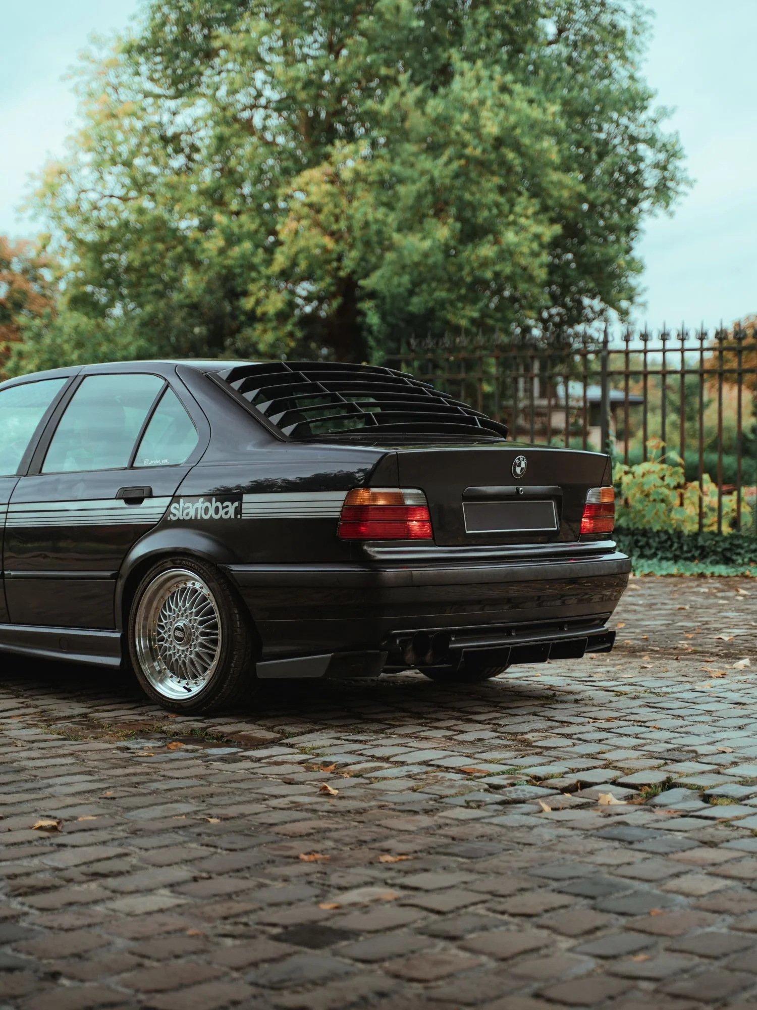 Voiture noire BMW classique stationnée sur une route en pavés, avec un arbre et une grille métallique en arrière-plan.