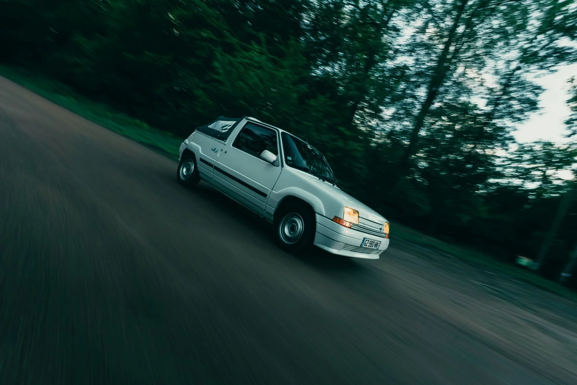 Une voiture blanche de style vintage conduit rapidement sur une route bordée d'arbres verts, la scène est légèrement floue à cause de la vitesse.