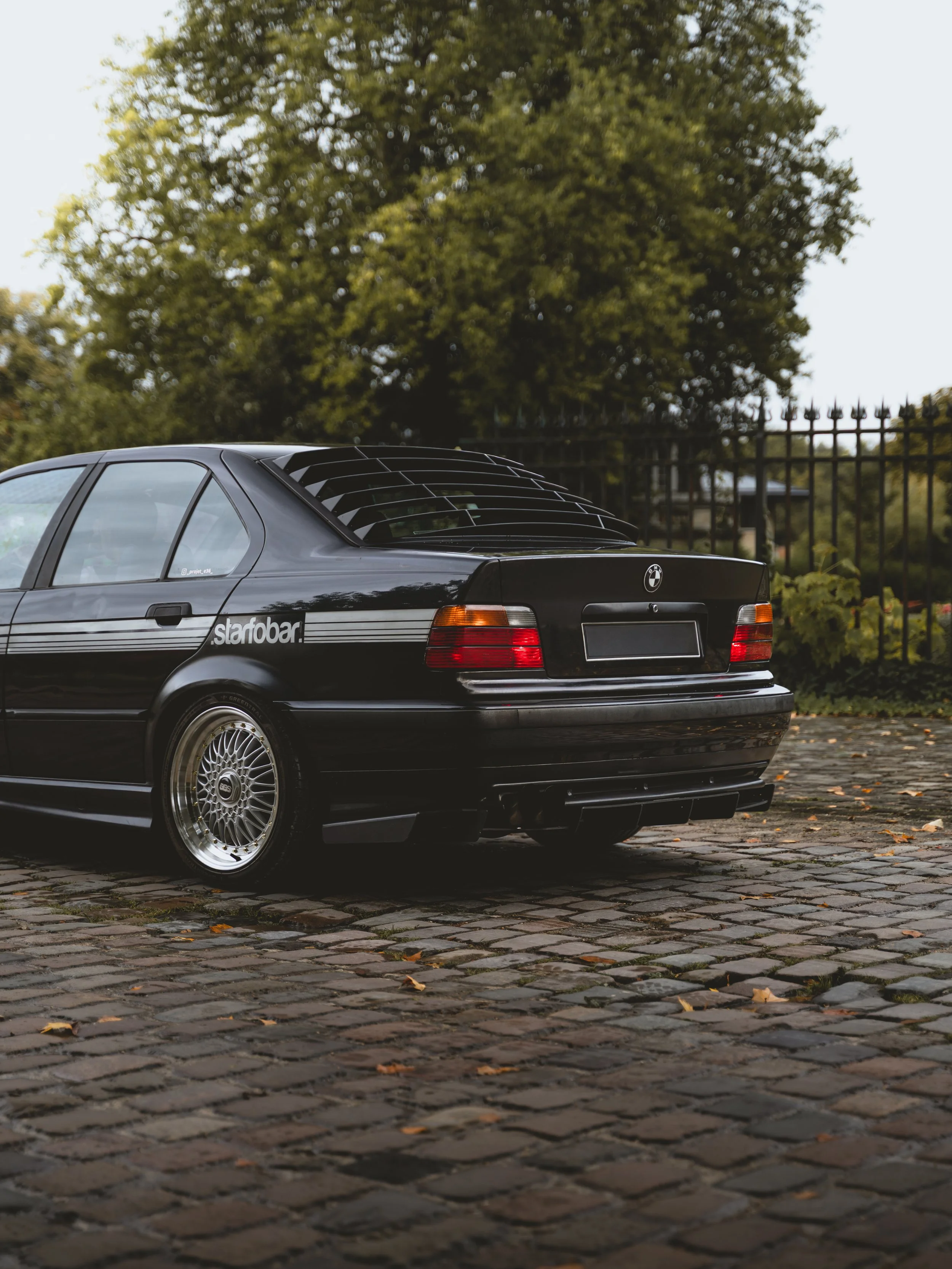 Voiture noire de style BMW ancienne, stationnée sur une chaussée en pavés, avec un arrière en vue et un arbre en arrière-plan.