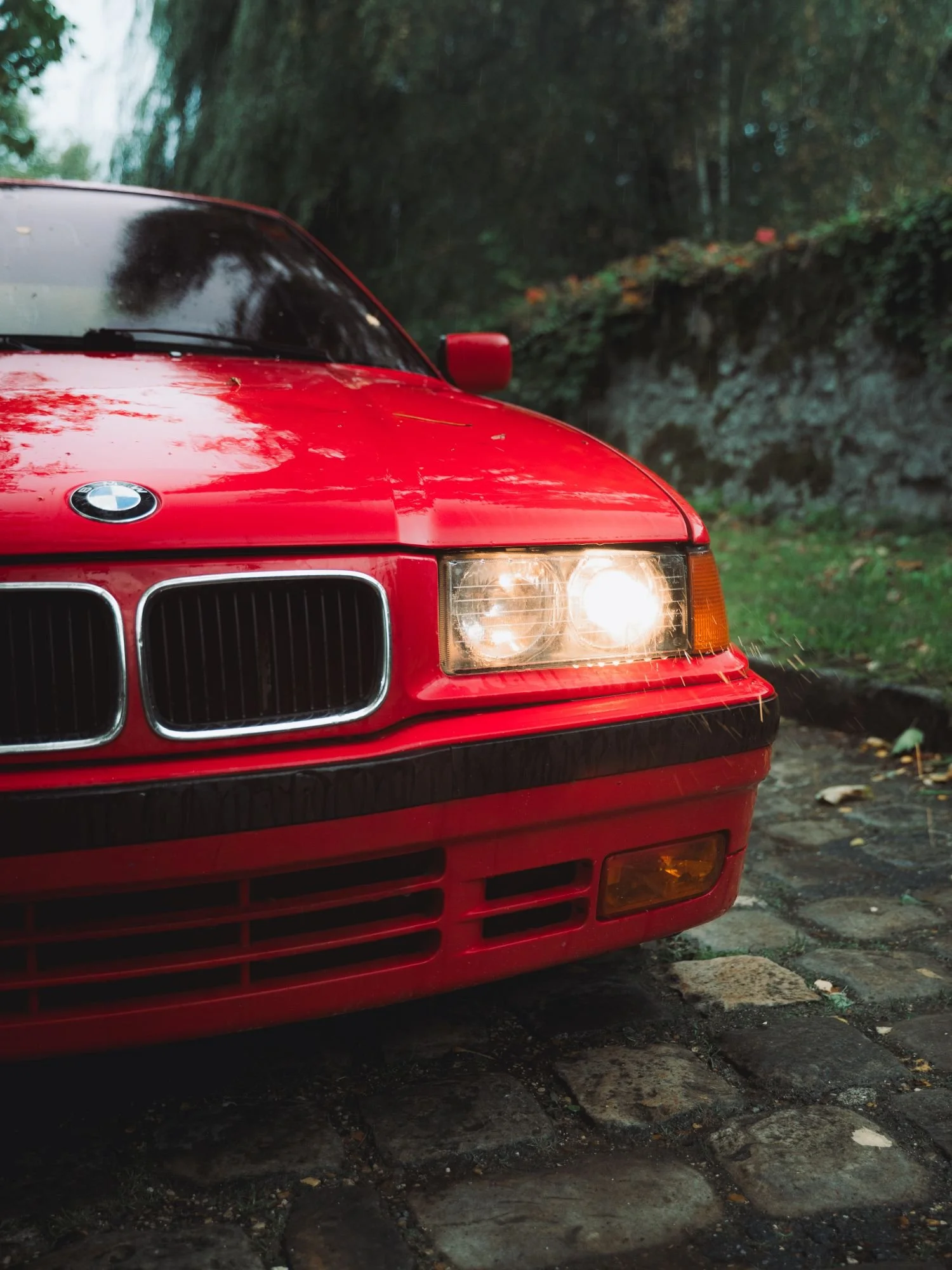 Voiture rouge BMW de face, stationnée sur un chemin en pierre sous des arbres, d'un côté de l'image