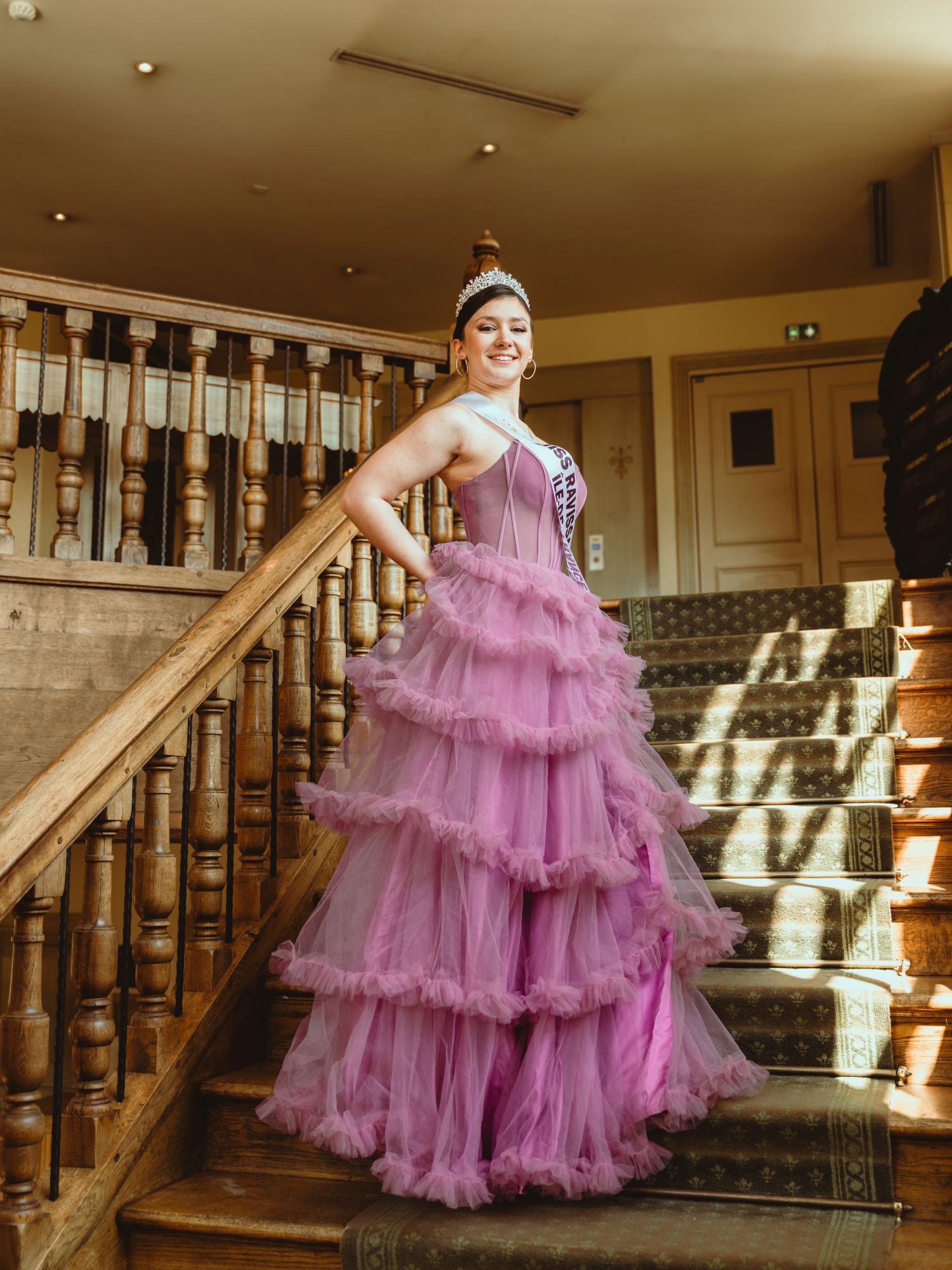Jeune femme portant une robe de soirée rose à volants, une couronne, un sash, et un diadème, sur un escalier en bois dans un intérieur.