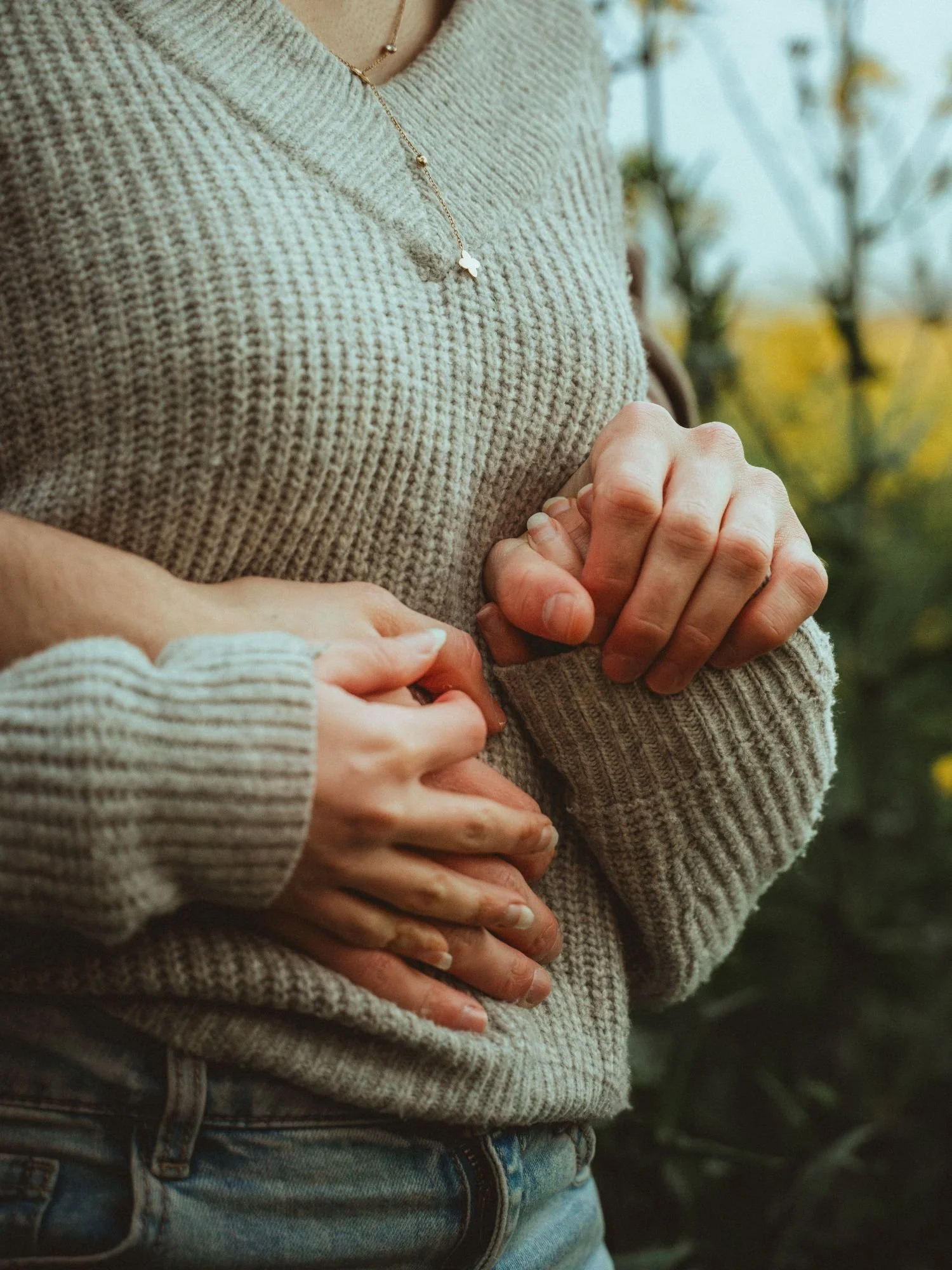 Deux personnes, une adulte et un enfant, tenant leur ventre, suggérant qu'elles attendent un bébé. La personne adulte porte un pull en laine beige et une chaîne, tandis que l'enfant porte un pull en laine gris clair. L'arrière-plan montre une nature 