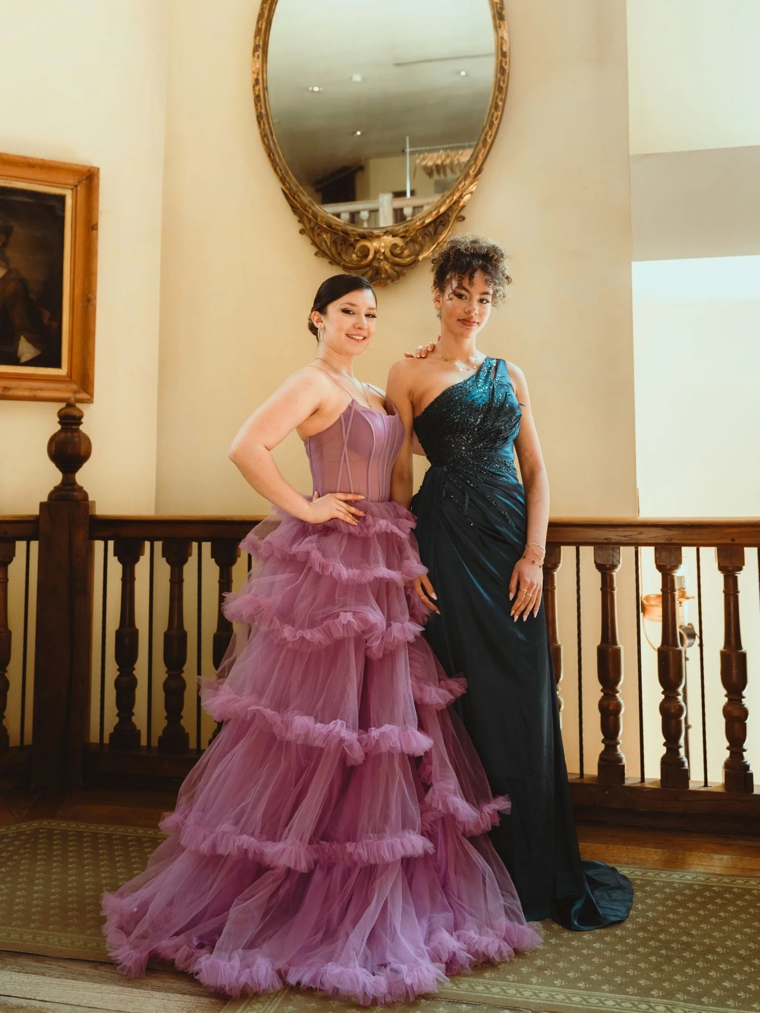 Deux femmes en robes de soirée élégantes posant sur un balcon intérieur, avec un miroir doré au mur derrière elles.