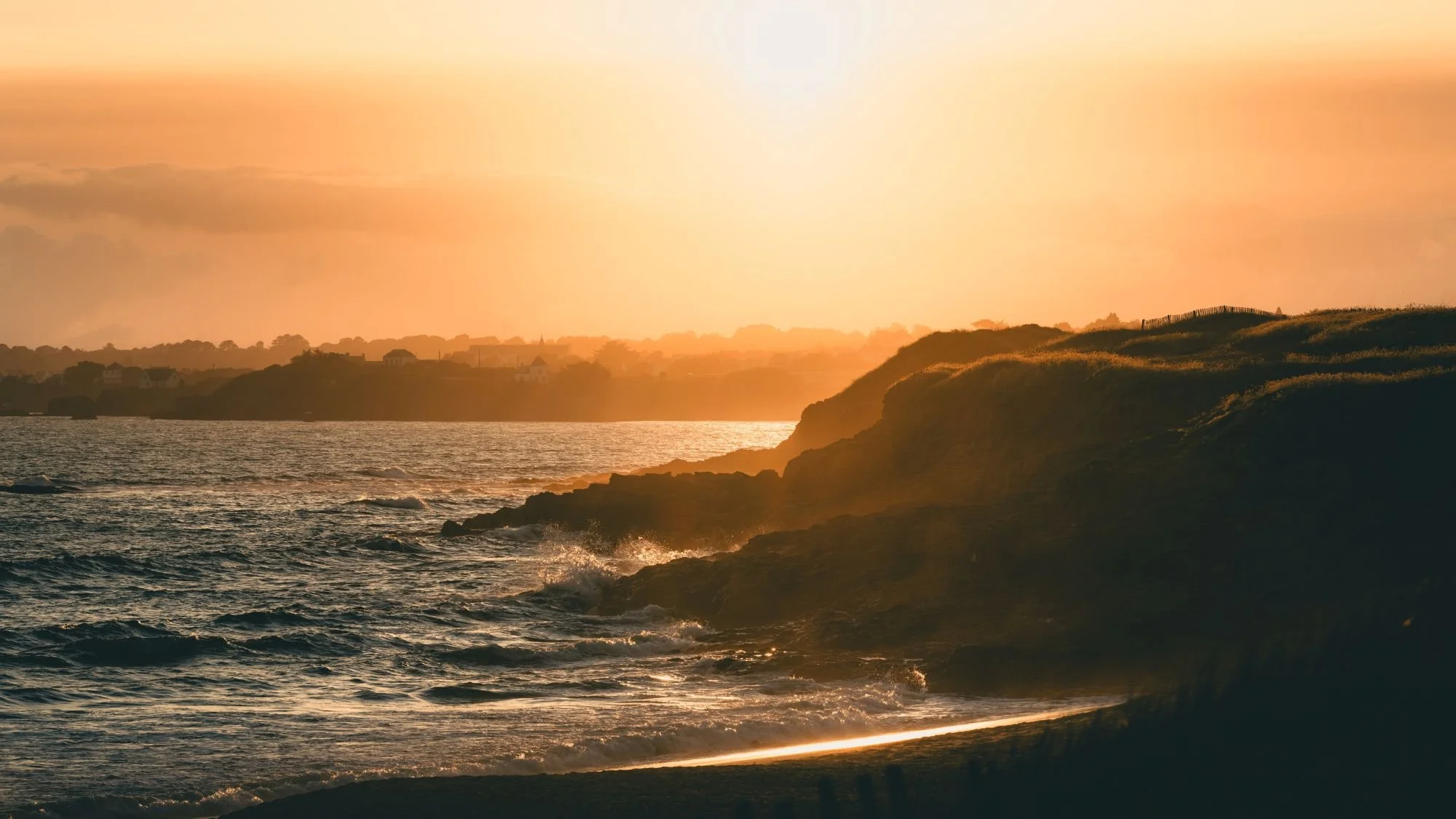 Coucher de soleil sur une côte rocheuse avec la mer en premier plan, horizon lointain et ciel orangé.
