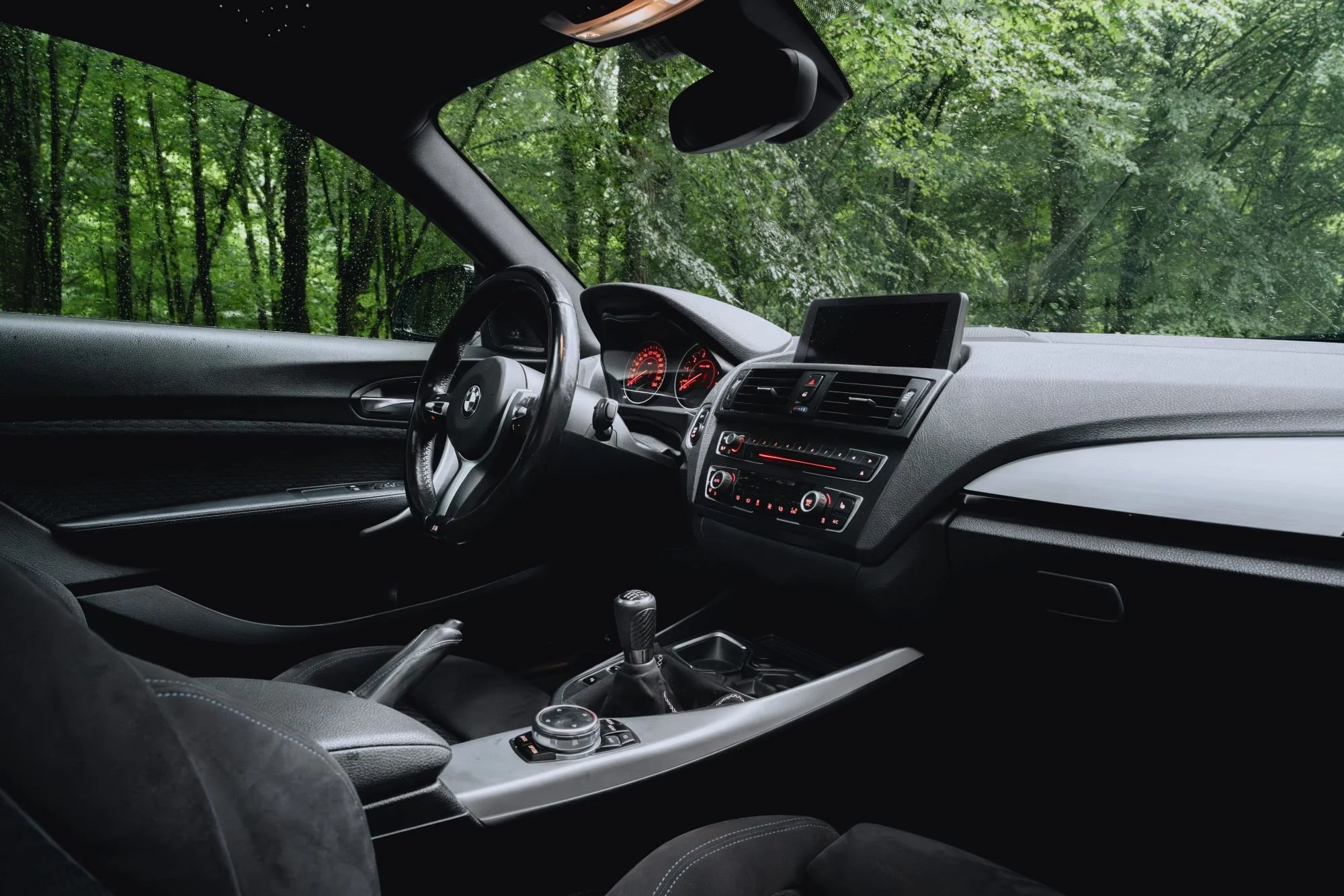 Intérieur d'une voiture avec tableau de bord, volant, et siège, vue à travers le pare-brise sur un paysage forestier humide.