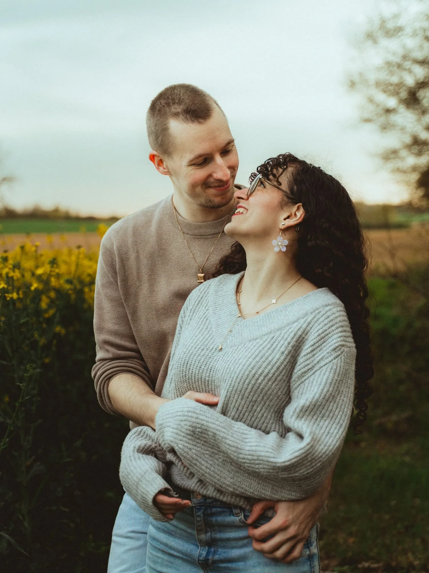 Un couple s'aime dans un champ, la femme avec des lunettes et des boucles d'oreilles blanches, le homme lui tient la taille. C'est une scène romantique en plein air.