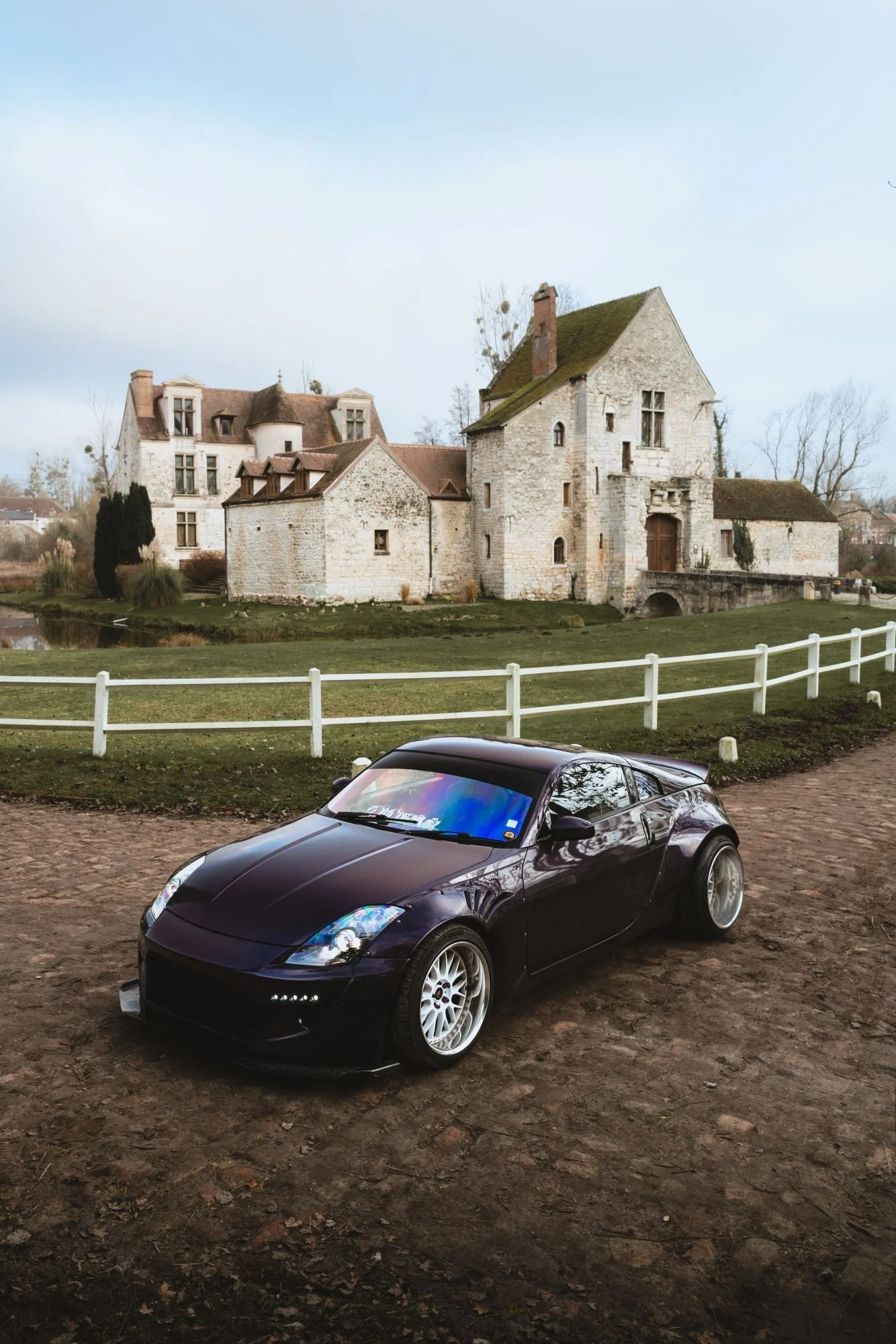 Voiture de sport noire parkée sur un chemin en terre devant un vieux château en pierre avec un toit en ardoise, sous un ciel partiellement nuageux.