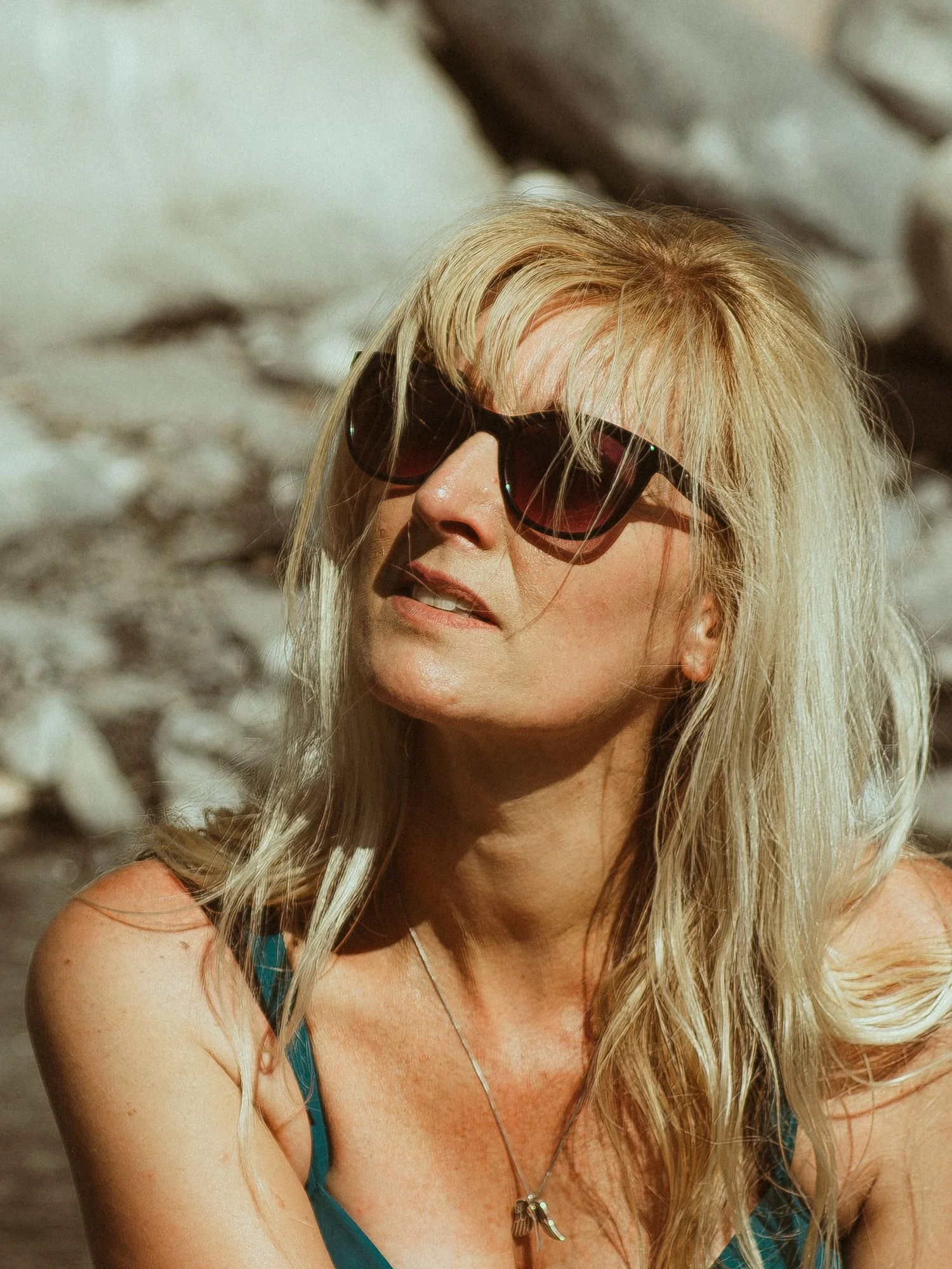 Une femme blonde portant des lunettes de soleil foncées et un haut bleu, posant en extérieur avec un fond rocheux.