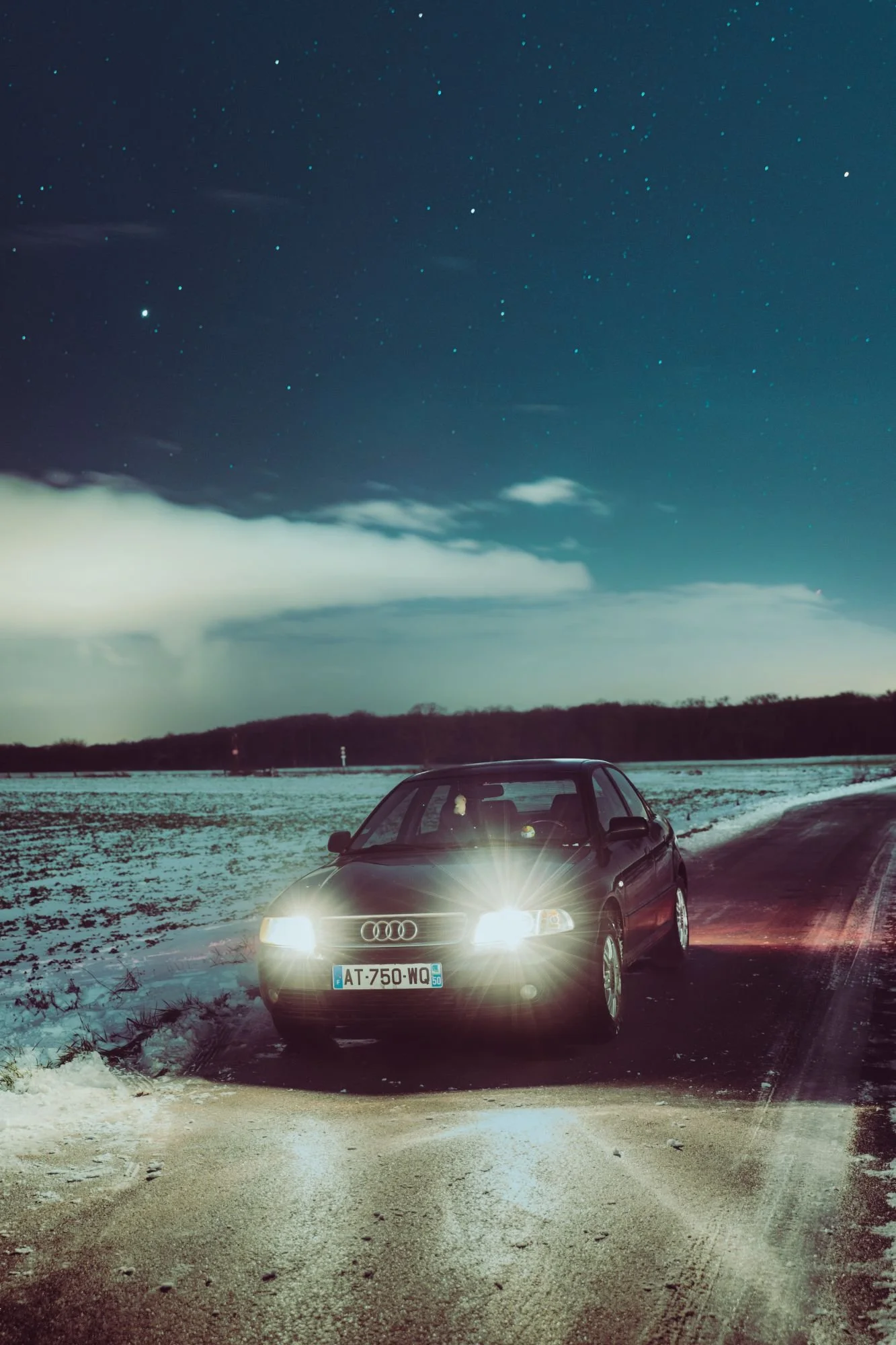 Une voiture noire Audi avec ses phares allumés sur une route enneigée la nuit, avec un ciel étoilé en arrière-plan.