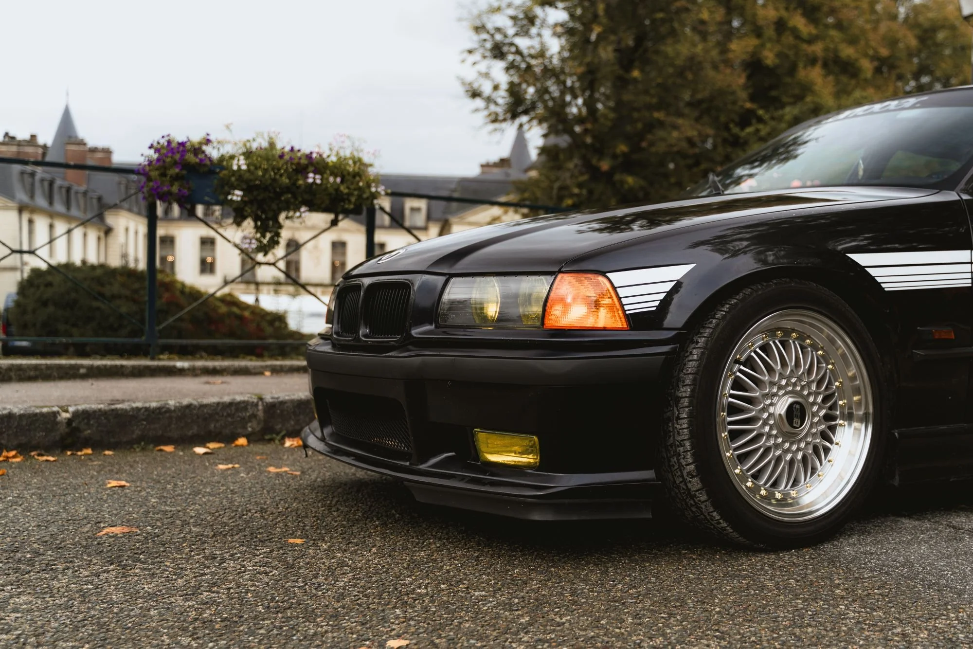 Voiture noire de sport BMW, vue partielle, stationnée sur une rue pavée avec des bâtiments anciens en arrière-plan, arbres avec feuilles d'automne, et des fleurs dans un pot suspendu.