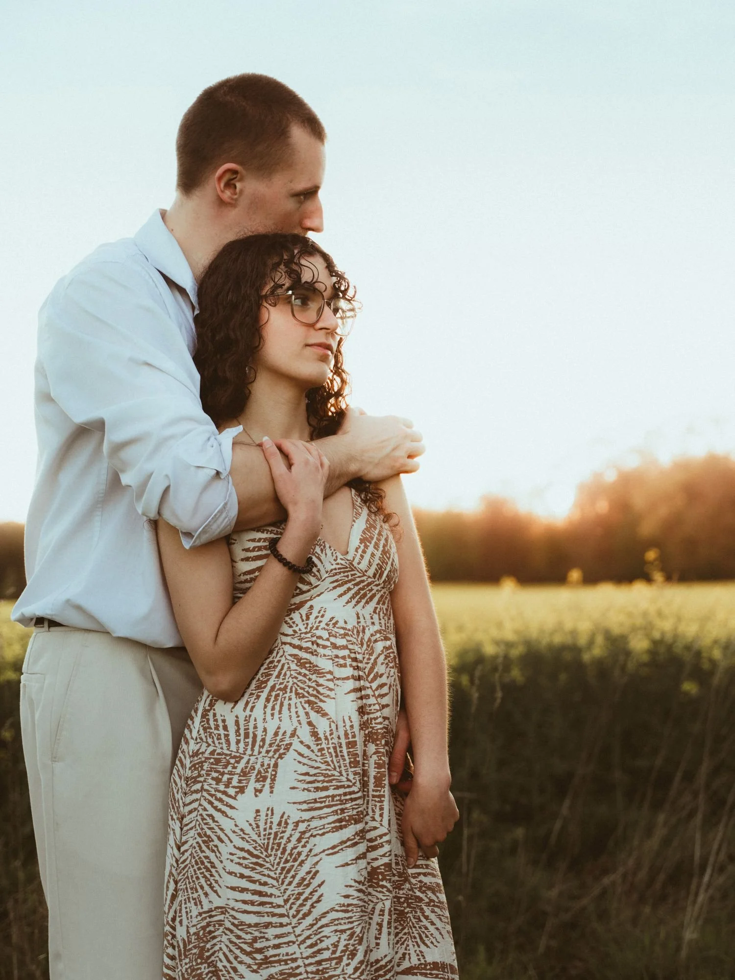Un homme et une femme debout dans un champ lors du coucher du soleil, l'homme embrassant la femme sur la tête.