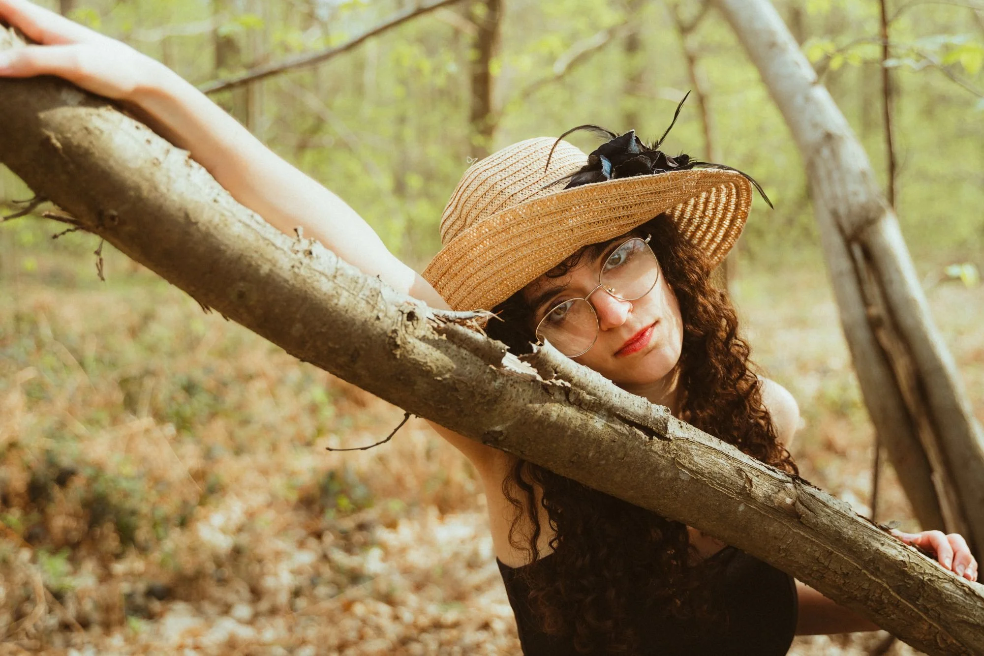 Une femme avec des cheveux bouclés portant un chapeau de paille et des lunettes, posant dans une forêt en s'appuyant sur une branche d'arbre.