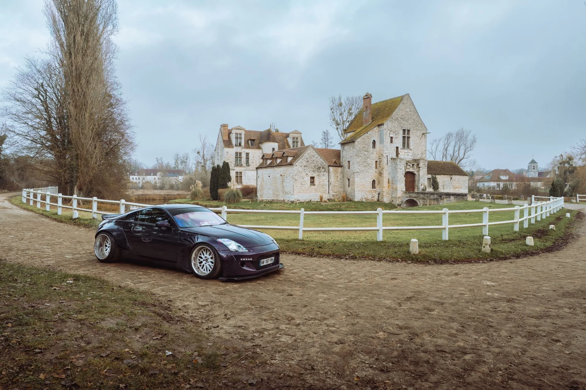Une voiture noire stationnée sur un chemin en terre devant un vieux château en pierre, avec des arbres et des bâtiments en arrière-plan.