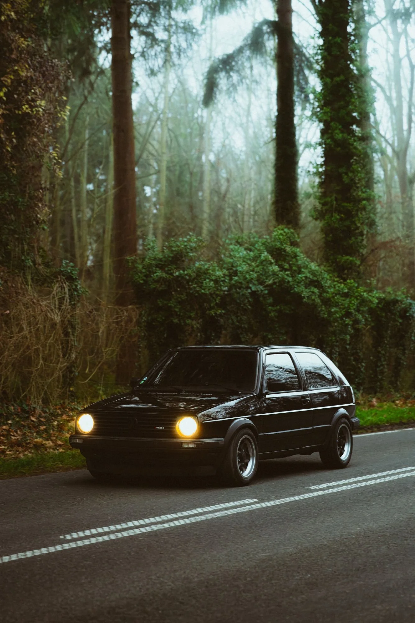 Une voiture noire garée sur une route en forêt, avec ses phares allumés, dans un environnement naturel verdoyant.