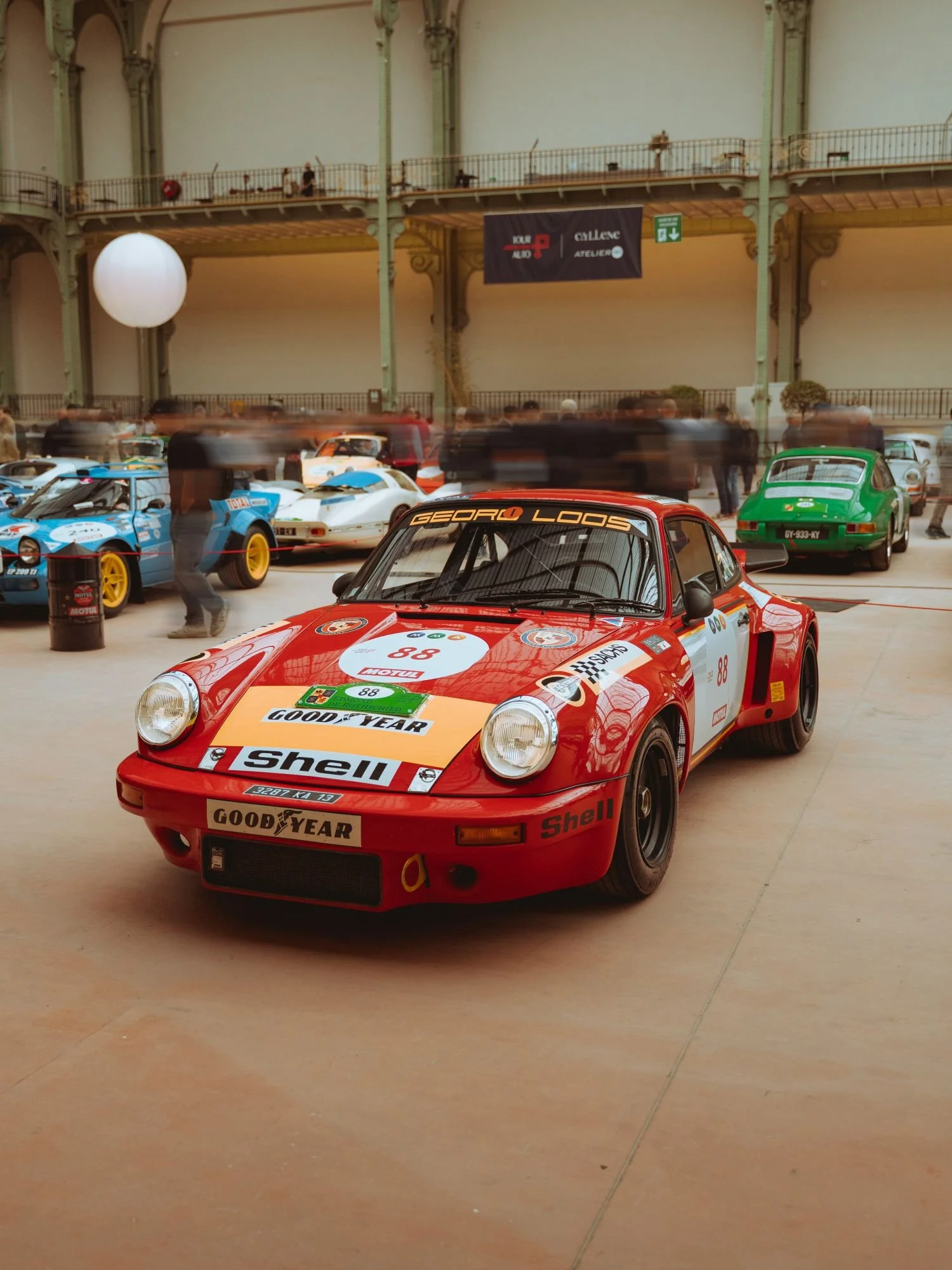 Une voiture de course Porsche rouge avec des logos Shell et Goodyear, exposée dans un bâtiment avec d'autres voitures anciennes et des spectateurs en arrière-plan.