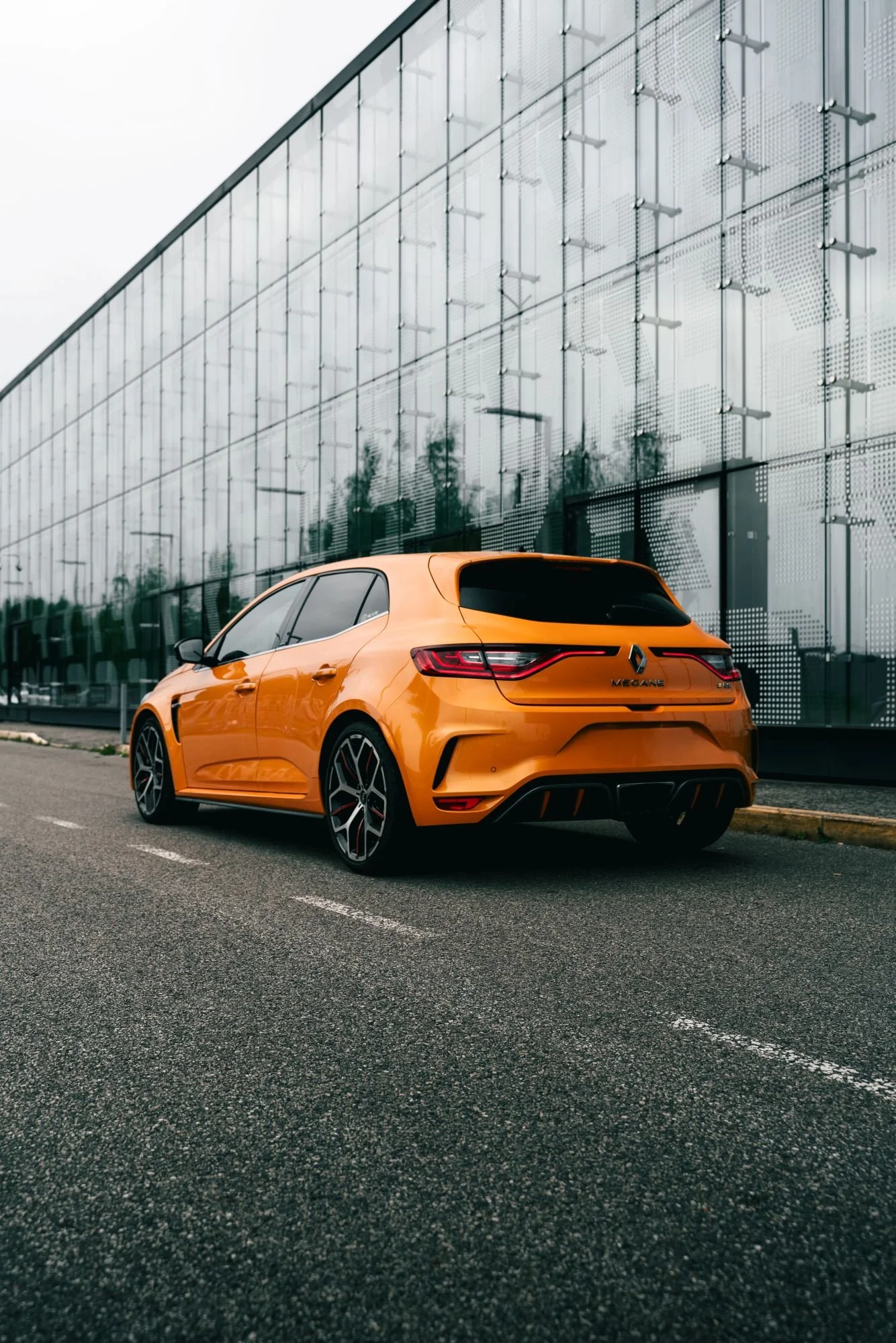 Une voiture orange Renault Megane Sport à l'arrêt sur une route, devant un bâtiment en verre moderne.