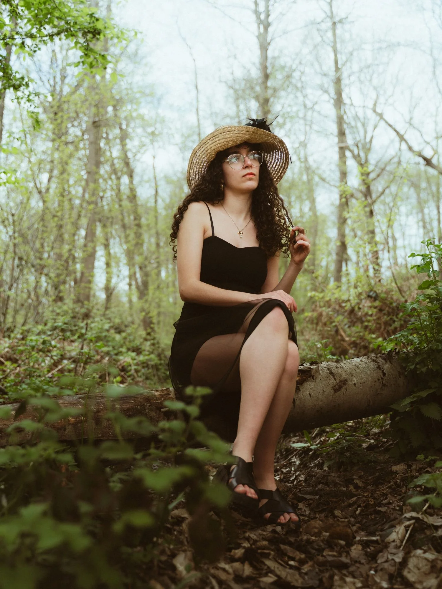 Jeune femme assise sur un tronc d'arbre dans une forêt, portant une robe noire, un chapeau en paille, des lunettes et des sandales noires.