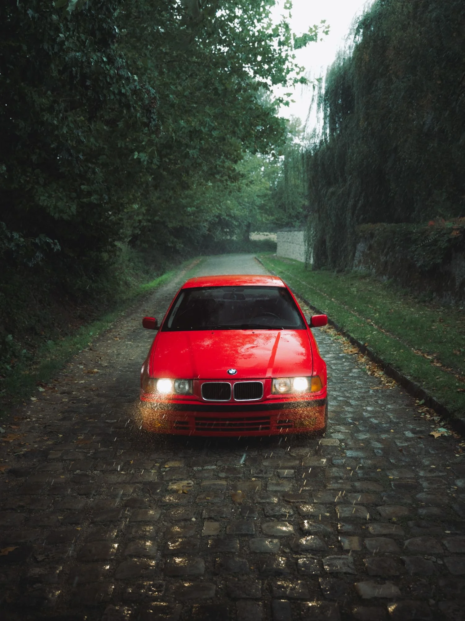 Voiture rouge sur une route pavée sous la pluie, entourée d'arbres verts et de haies.