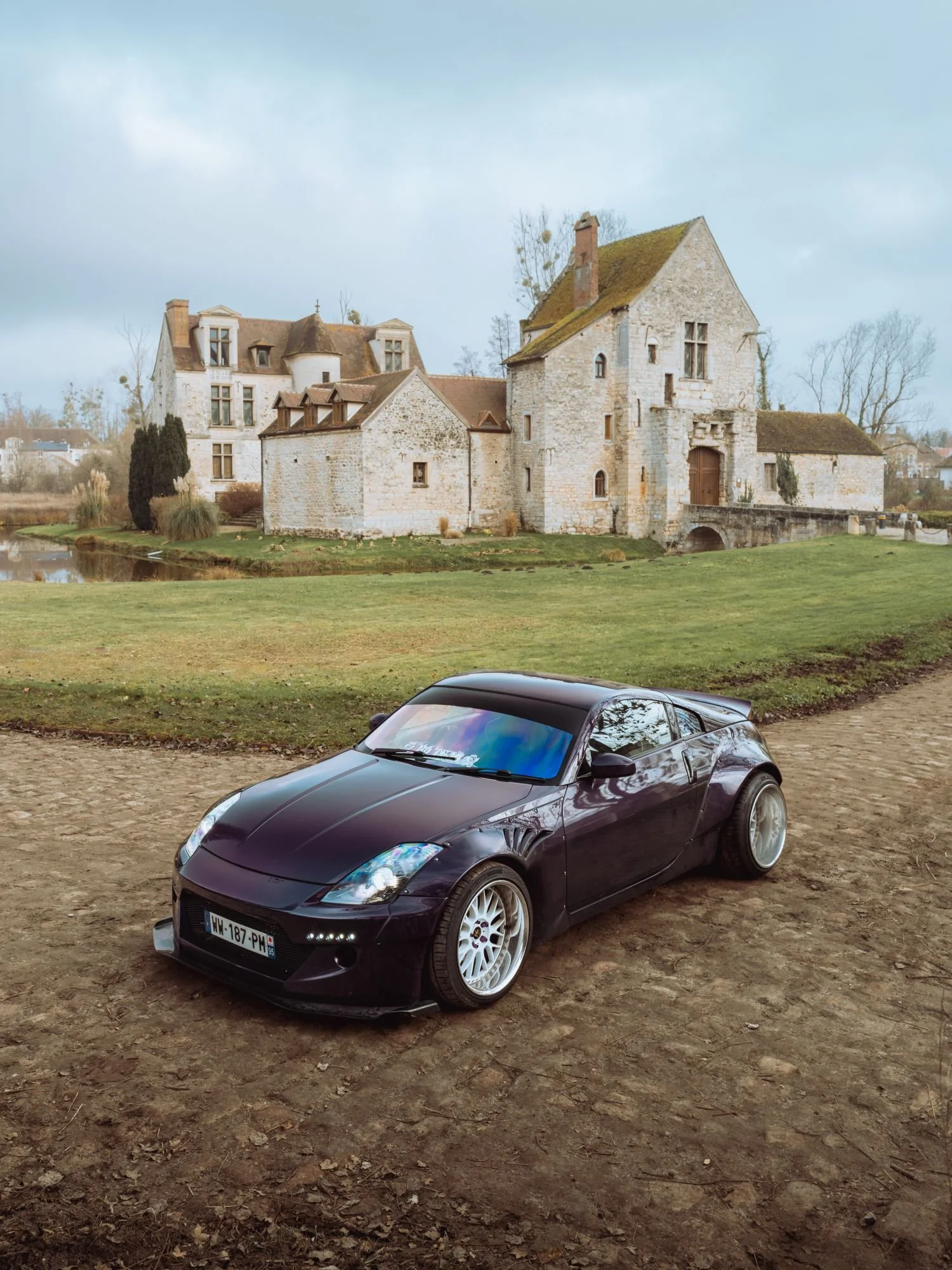 Voiture de sport noire garée sur un terrain en terre devant un vieux château en pierre avec un toit en tuiles et un paysage rural en arrière-plan.