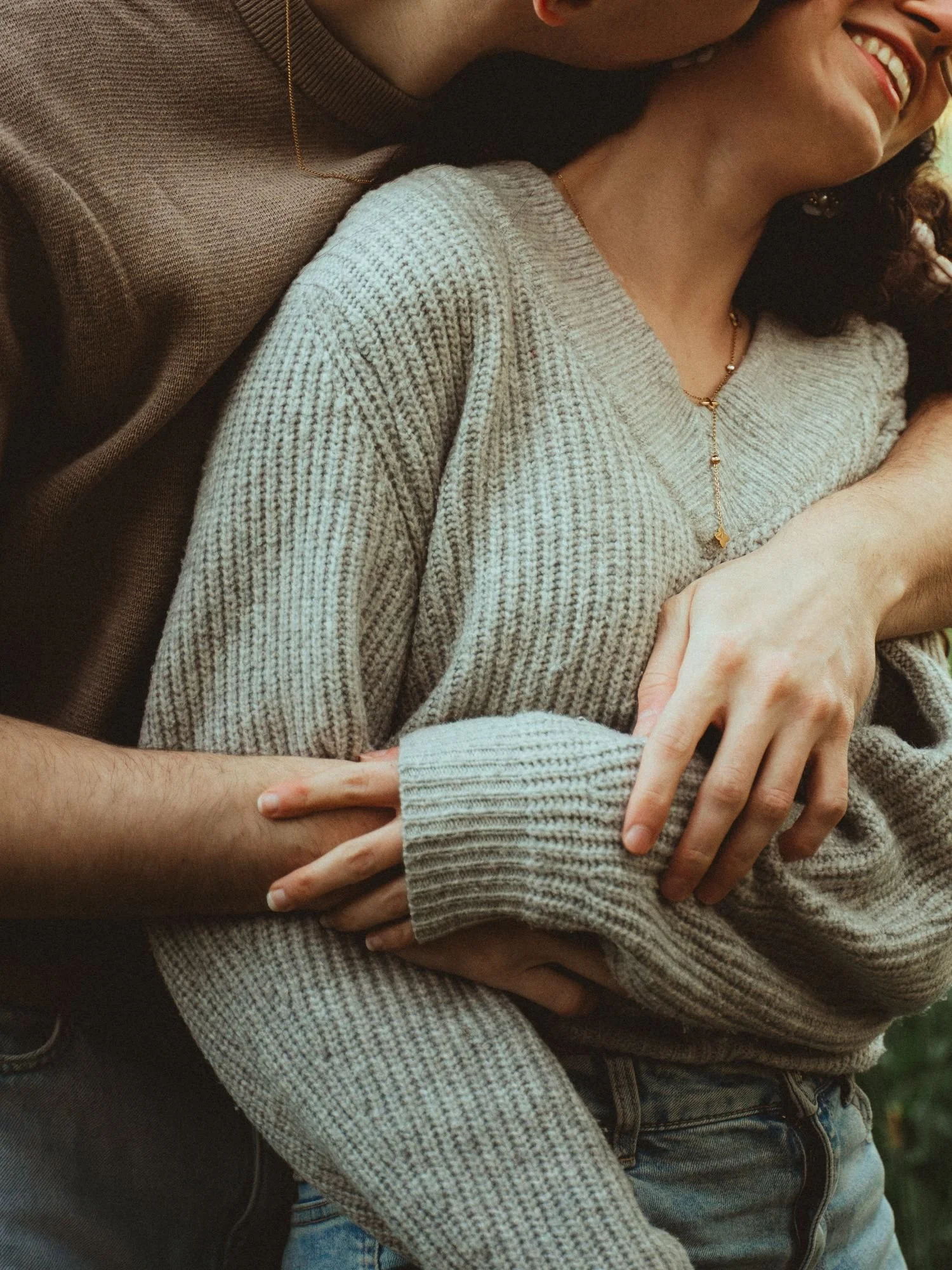Une personne souriante porte un pull gris clair et une chaîne en or, l'autre personne a le bras autour de la première, leur main est visible.