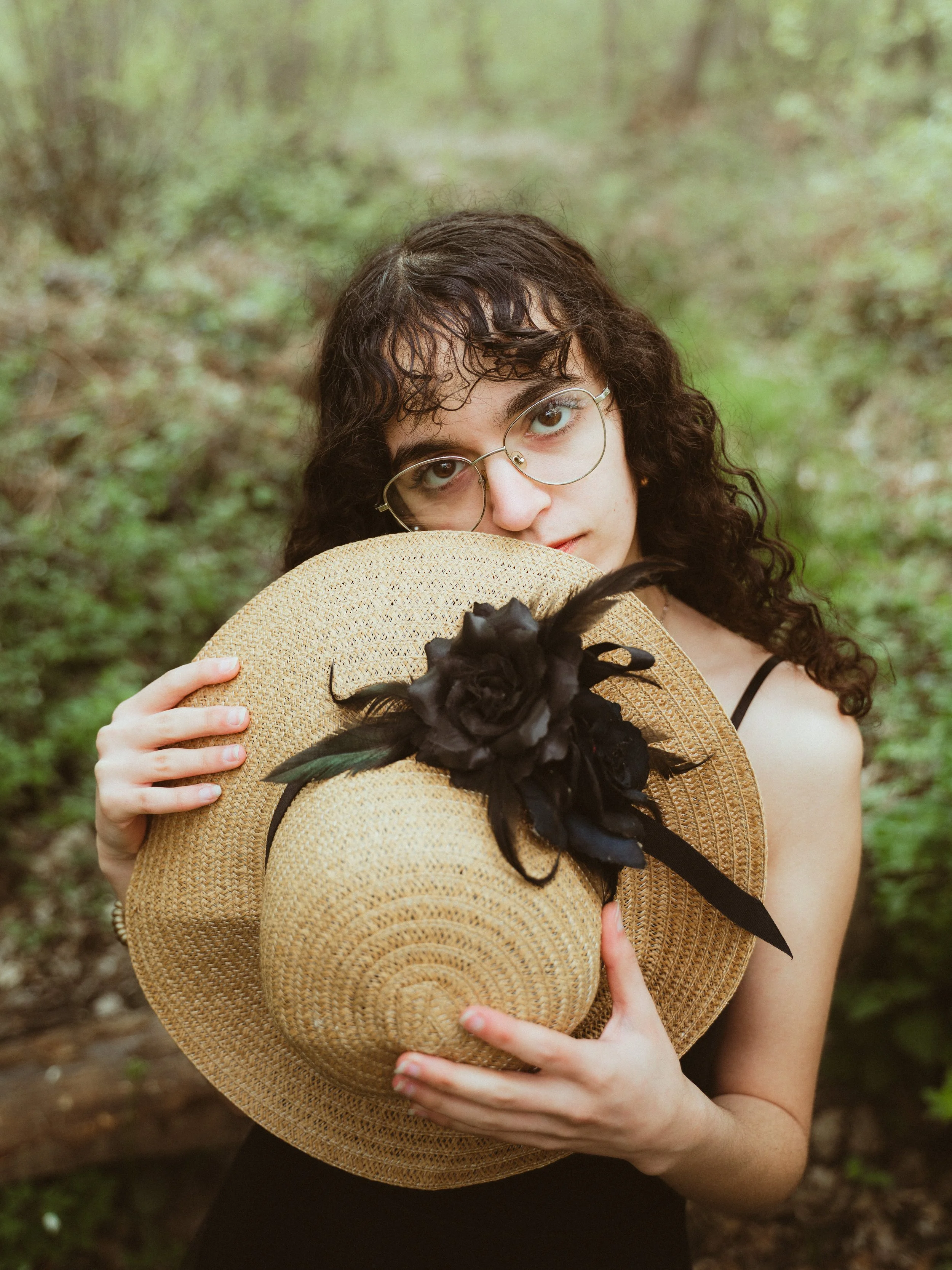 Jeune femme aux cheveux bouclés, portant des lunettes, tenant un chapeau de paille avec une décoration de fleur noire dans un environnement forestier.