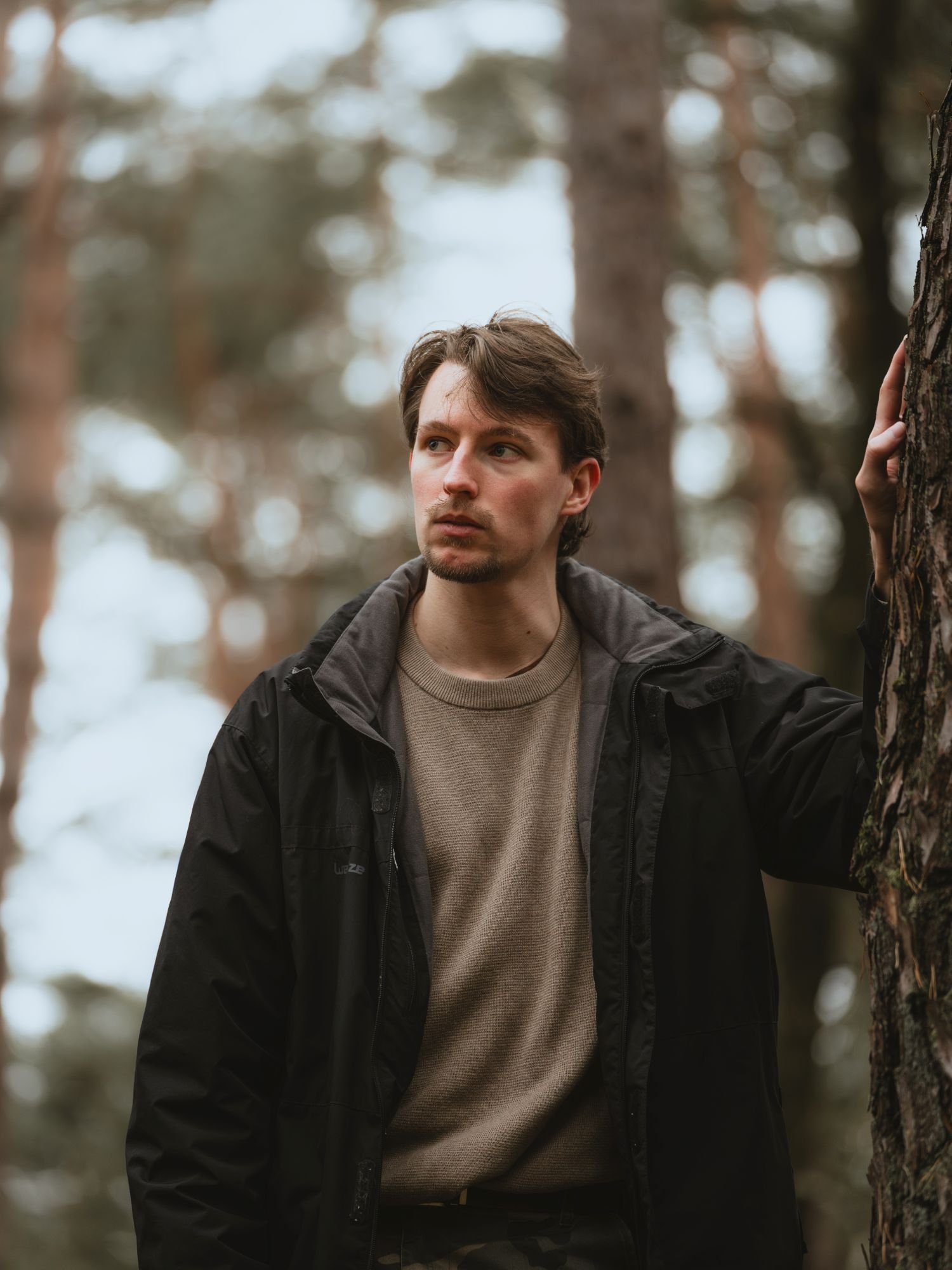 Un jeune homme avec des cheveux bruns, portant une veste noire et un pull beige, se tient dans une forêt, appuyé contre un arbre.