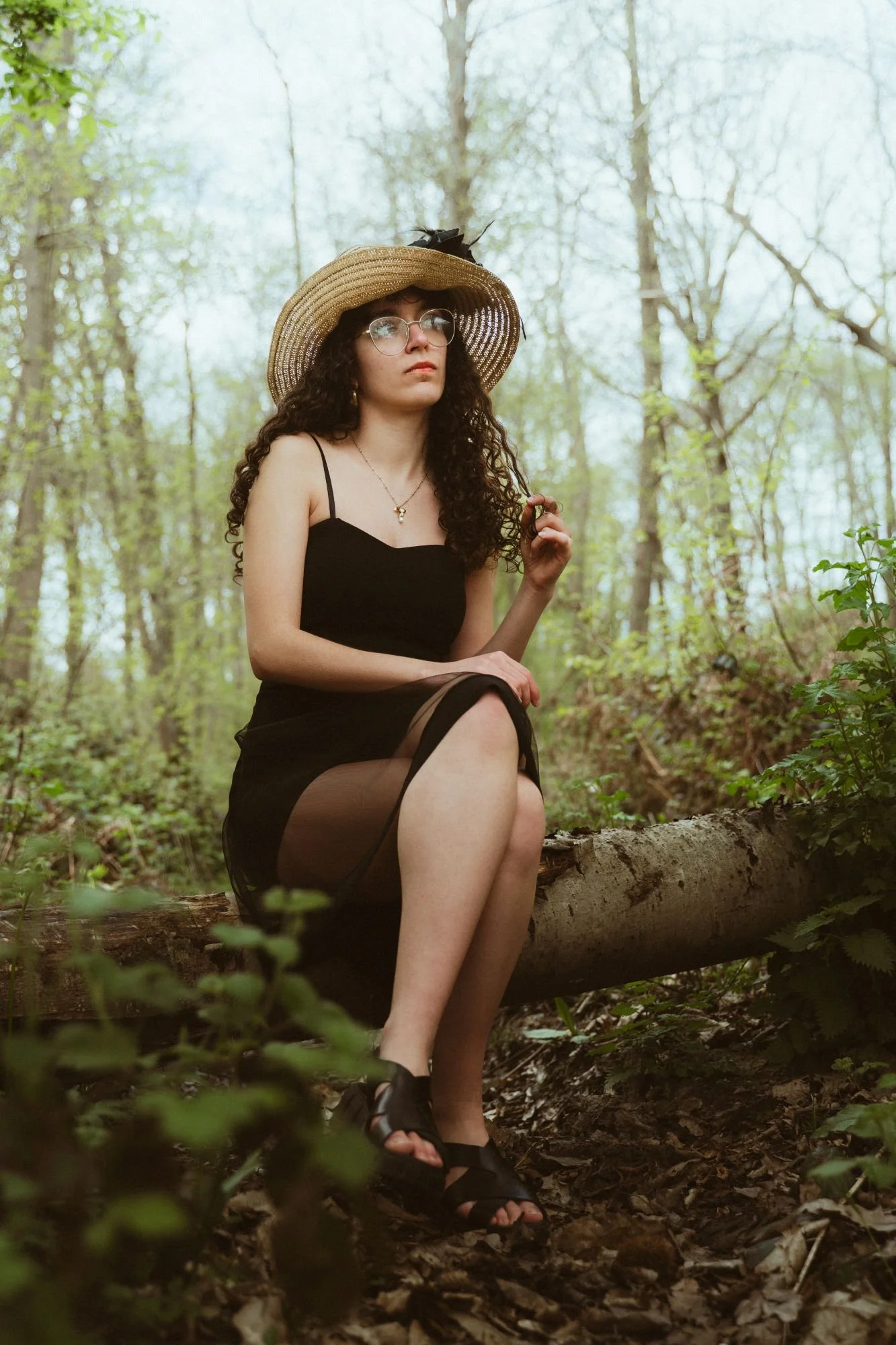 Une jeune femme assise sur un tronc dans une forêt, portant un chapeau en paille, une robe noire, des lunettes et des sandales, avec un ciel nuageux en arrière-plan.