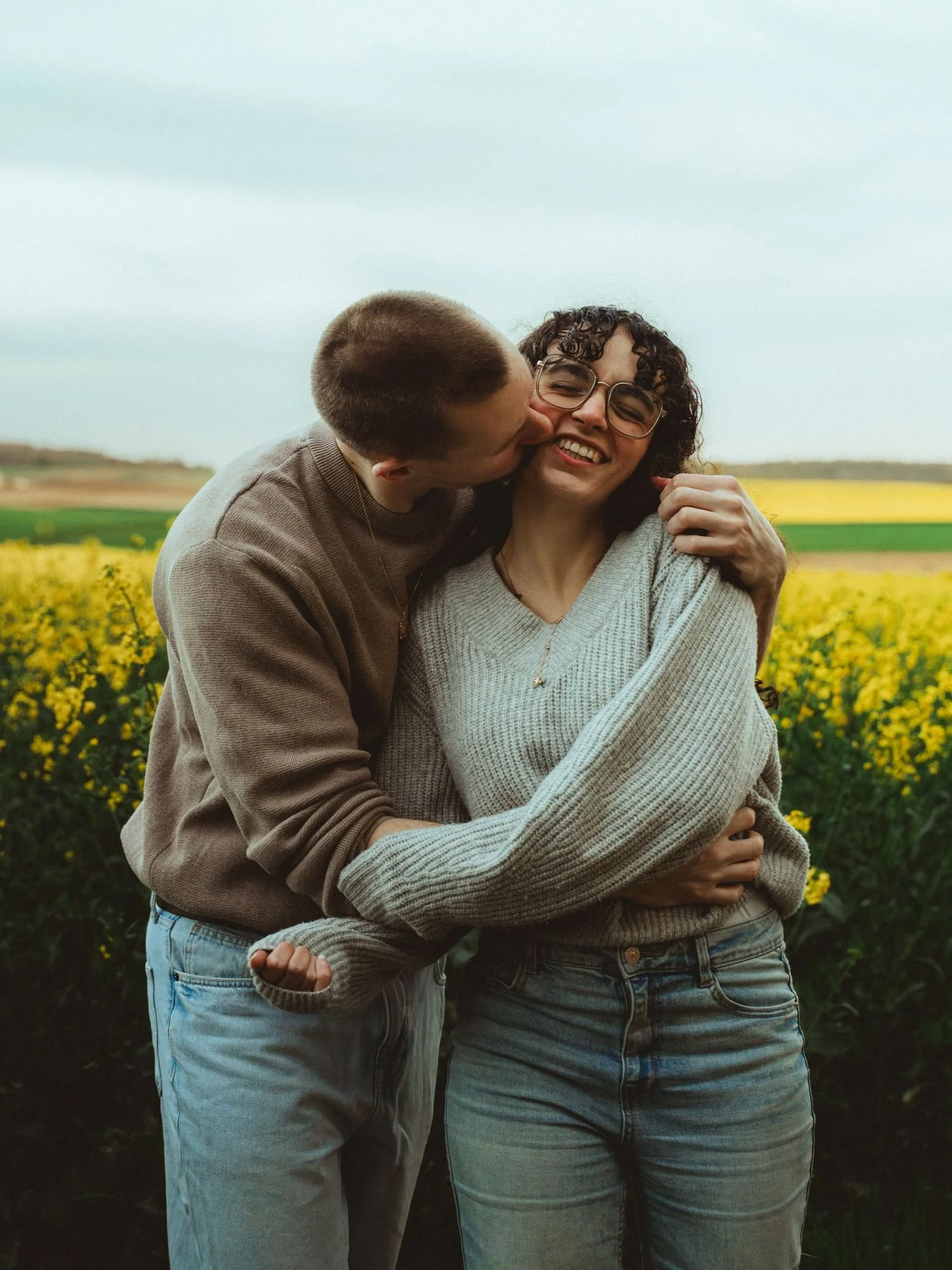 Un couple s'embrasse et rit dans un champ de fleurs jaunes, avec un ciel nuageux en arrière-plan.