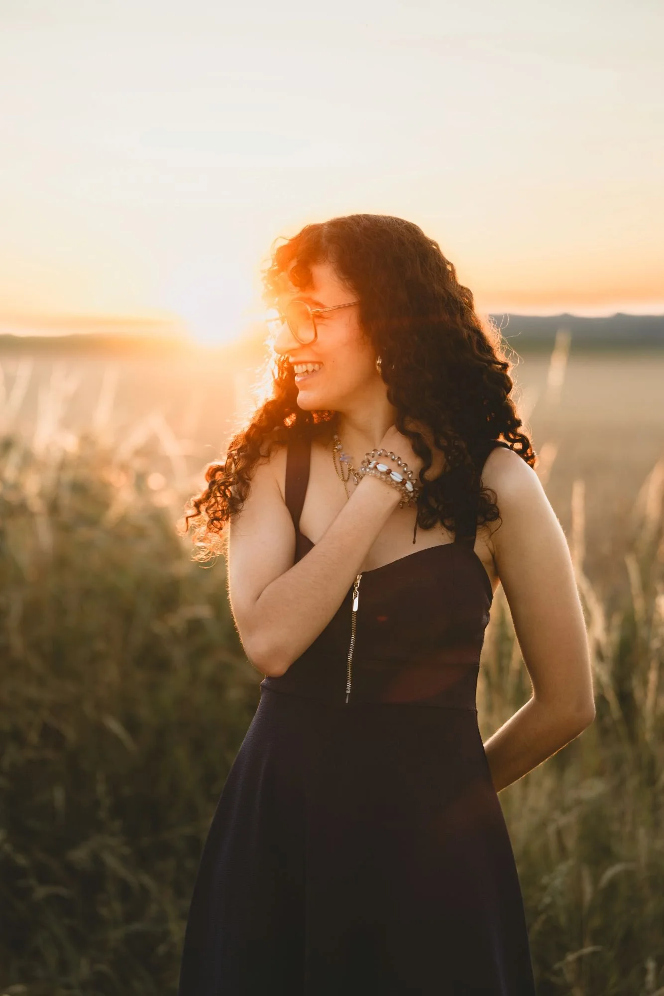 Une femme à cheveux bouclés, portant une robe noire, sourit dans un champ lors d'un coucher de soleil.