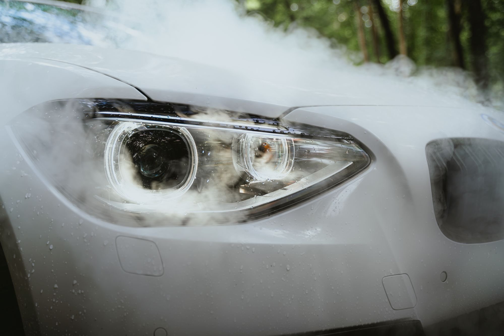 Photo d'une voiture blanche avec des phares modernes. Il y a de la vapeur ou de la fumée autour de la voiture, et on peut voir une forêt en arrière-plan.