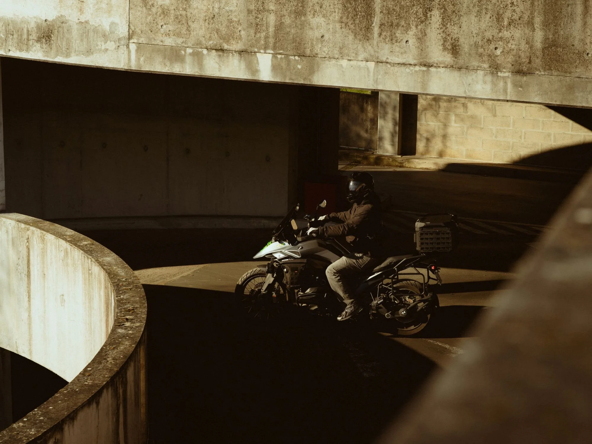 Un motard portant un casque noir et une veste marron, roulant dans un tunnel en béton avec des murs en brique.