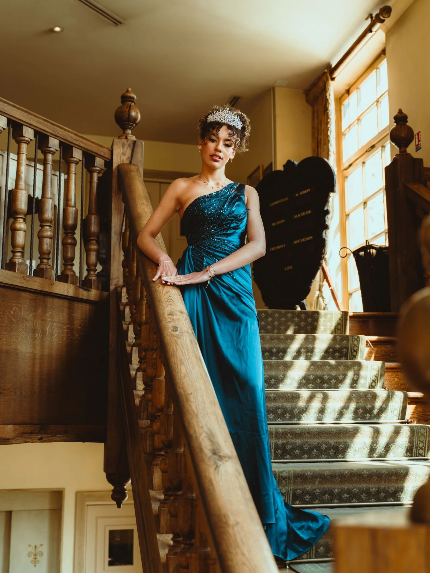 Une jeune femme en robe de soirée bleue sur un escalier en bois, dans un intérieur lumineux avec de grandes fenêtres.