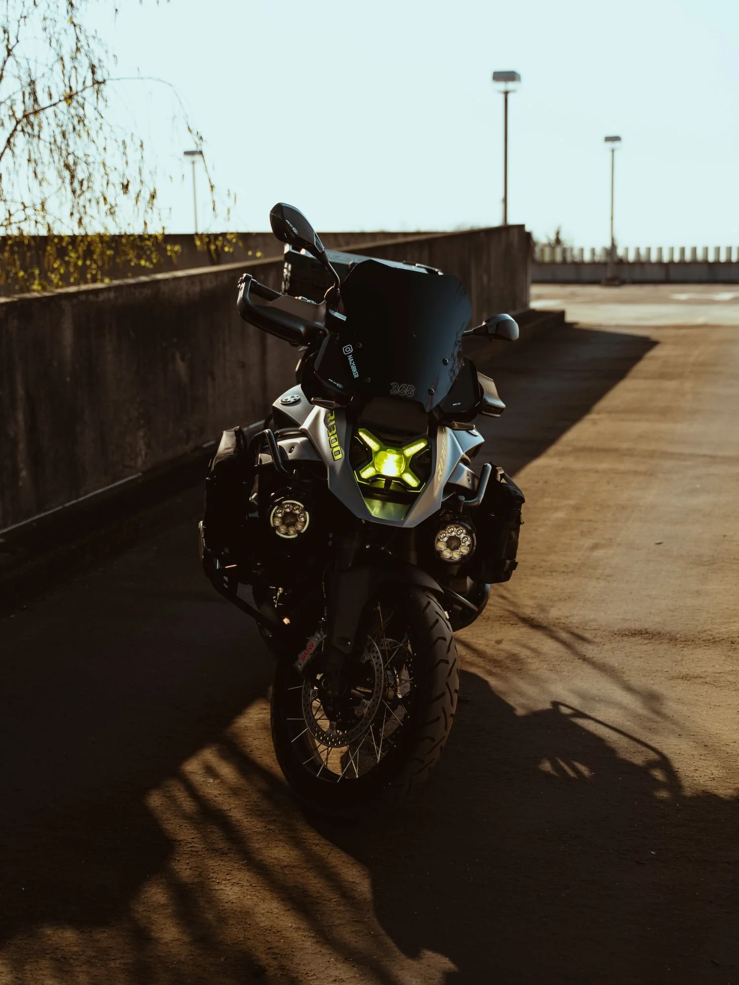 Une moto noire avec un pare-brise haut, stationnée sur un parking en plein air, avec un fond de ciel clair et de lampadaires.