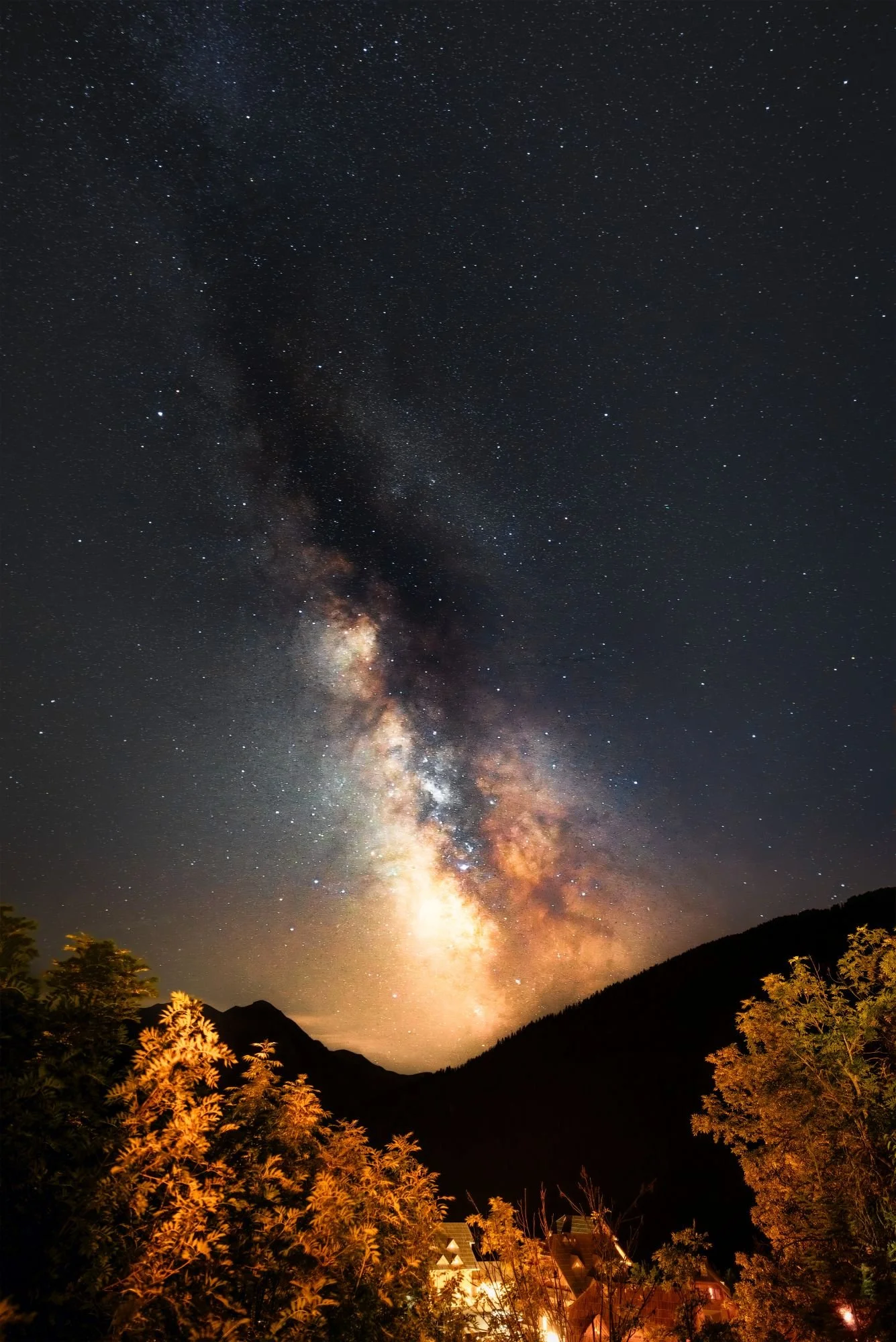 Ciel étoilé prominent avec la Voie lactée visible au-dessus de montagnes et de forêts, avec quelques maisons éclairées en bas.