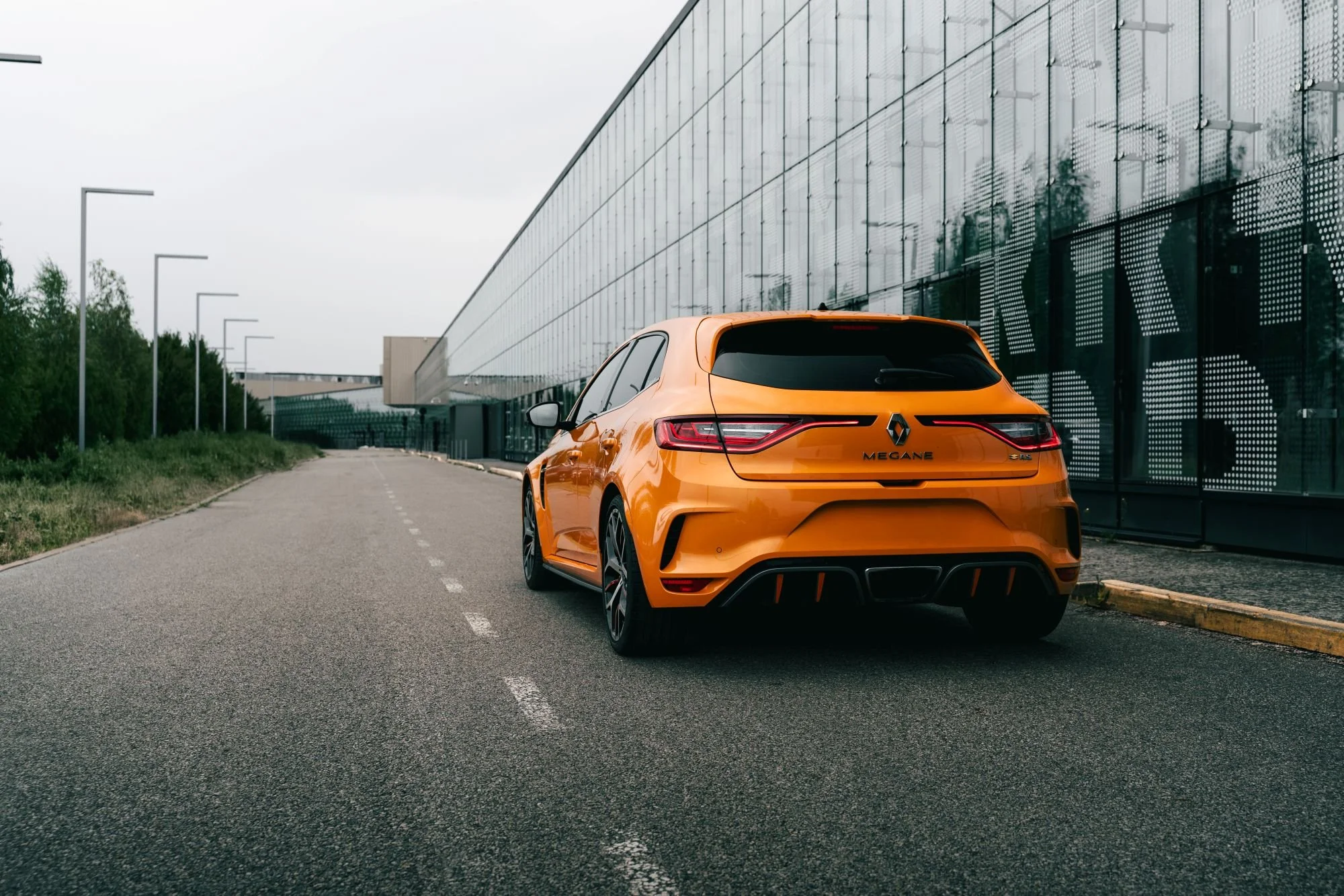 Voiture de sport orange garée le long d’un bâtiment moderne en verre avec des lampadaires alignés sur le côté gauche de la route.