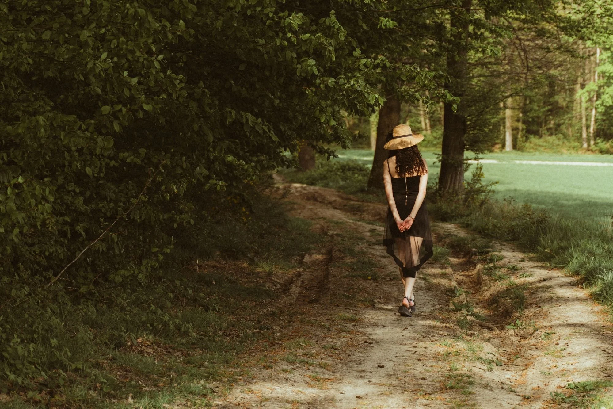 Une femme en robe noire, portant un chapeau de paille, marche seul sur un sentier forestier.
