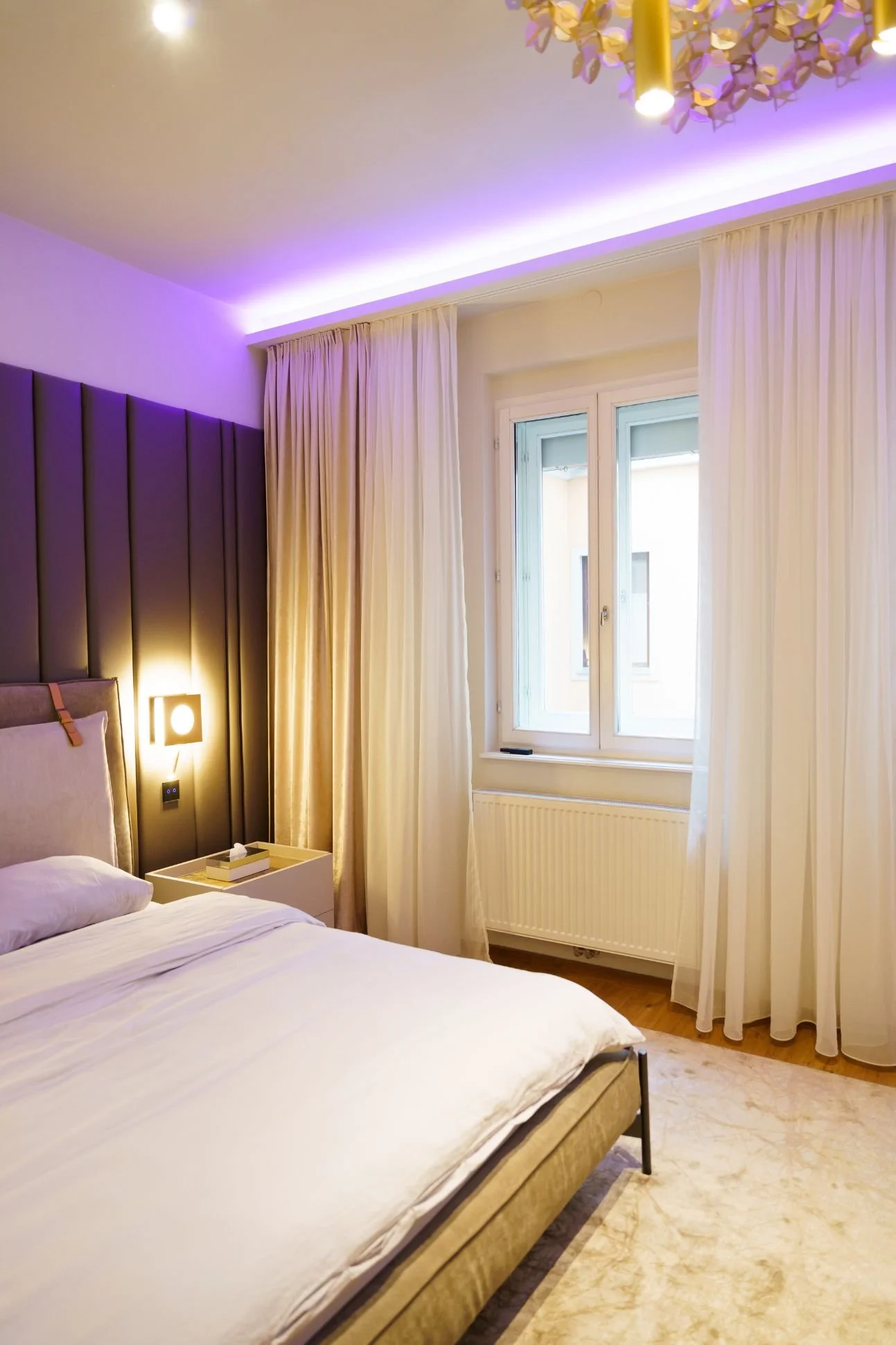 Modernes Schlafzimmer im Luxus- und Hotelstil mit gepolstertem Kopfteil, indirekter LED-Deckenbeleuchtung, hellen Vorhängen, maßangefertigtem Teppich, warmem Licht und Designer-Leuchten
