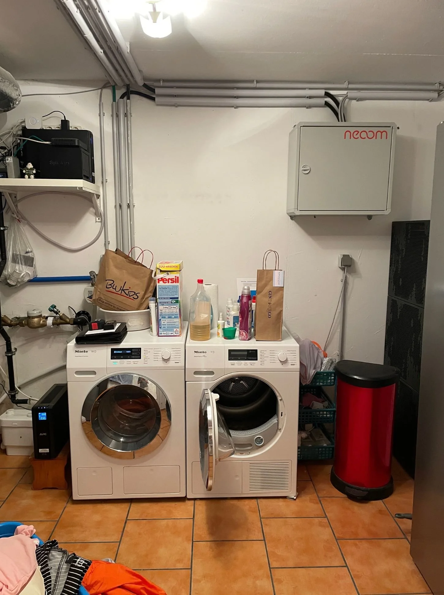 Waschmaschine, Trockner, Regale mit Reinigungsmitteln, Papiertüchern, Papierrollen, Kunststoffflaschen, Papiertragetaschen, ein schwarzer Wasserenthärter, ein roter Müllbehälter, ein Weißer Raumkasten mit roten Aufschrift, eine Geräte-Box, ein Hosenbügelsystem, ein schwarzer Müllbehälter, ein schwarzes Regal, eine Waschmaschine und einen Wäschetrockner, alles in einem Kellerraum mit terrakottafarbenen Fliesen.